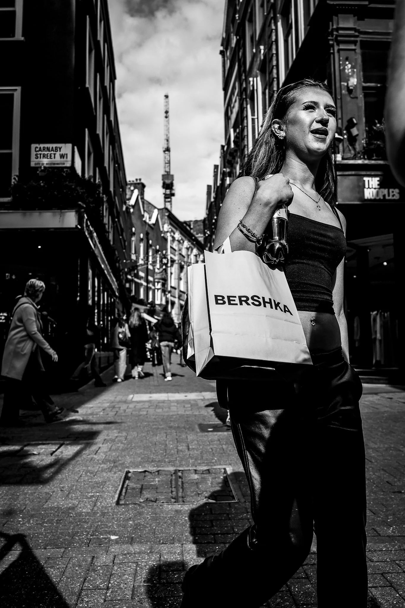 A woman with long hair is walking in an urban street, carrying a shopping bag with the brand name 'Bershka'. The street has buildings on both sides, a sign indicating 'Carnaby Street W1', and a crane in the background. The image is in black and white