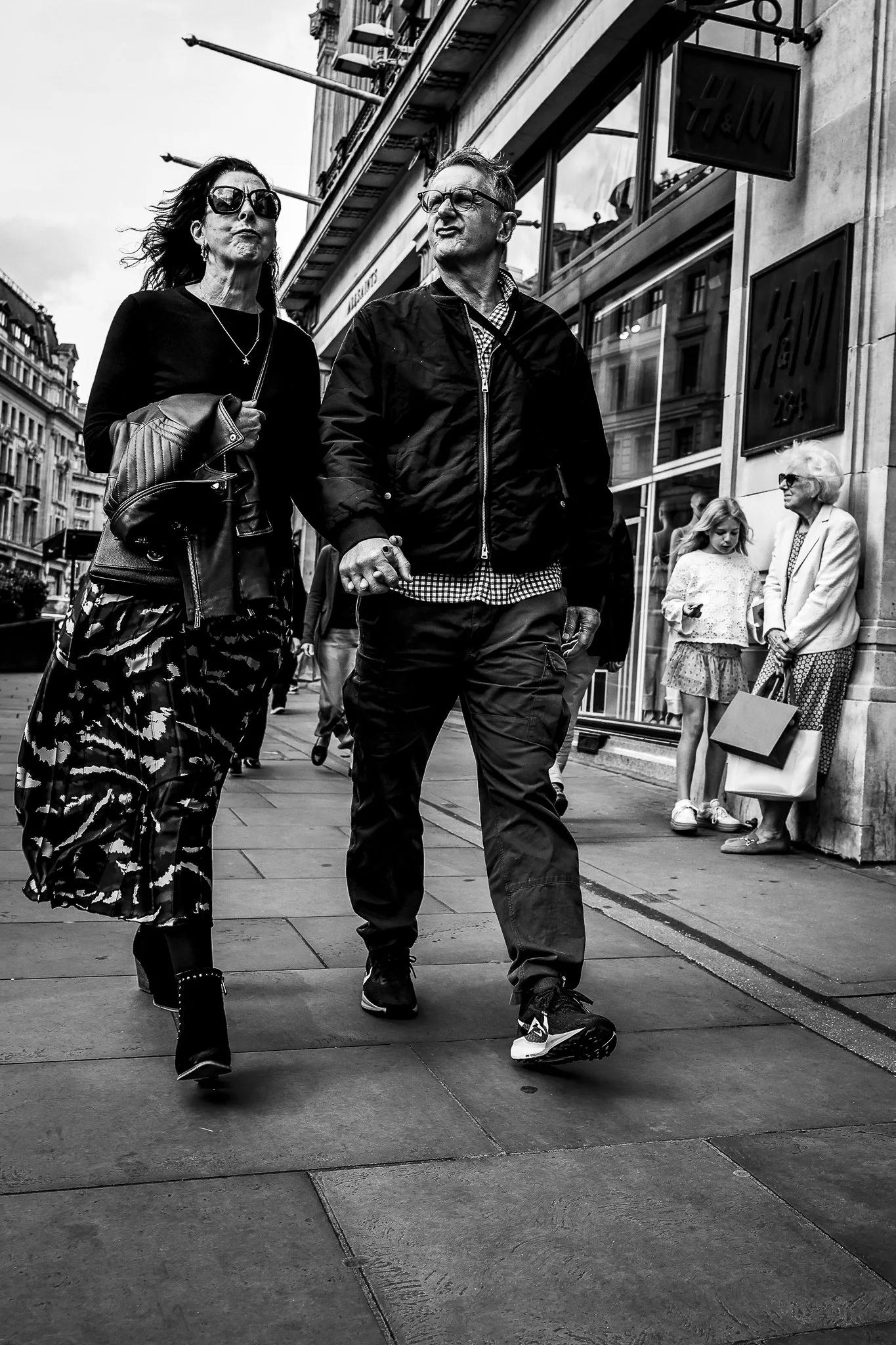 A black and white photo of a man and woman walking hand in hand on a city sidewalk. The woman wears large sunglasses, a dark top, and a floral skirt, carrying a purse. The man wears glasses, a jacket, and sneakers. In the background, there are other 