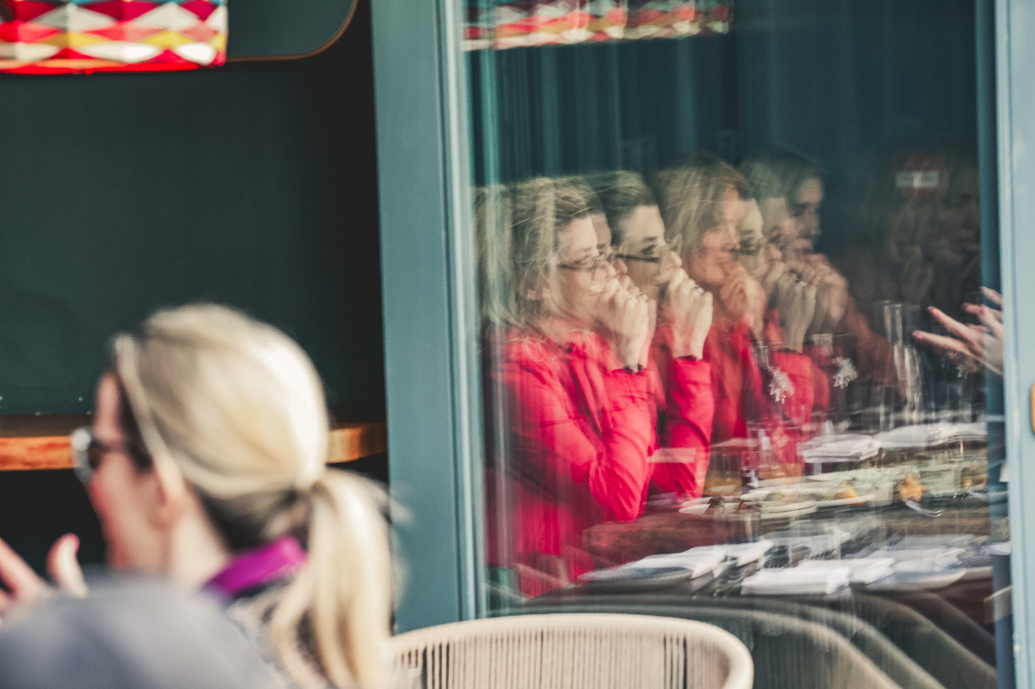 A reflection of four women wearing red shirts and glasses in a glass display case, with the women seated at a table or counter and blurred figures in the foreground.