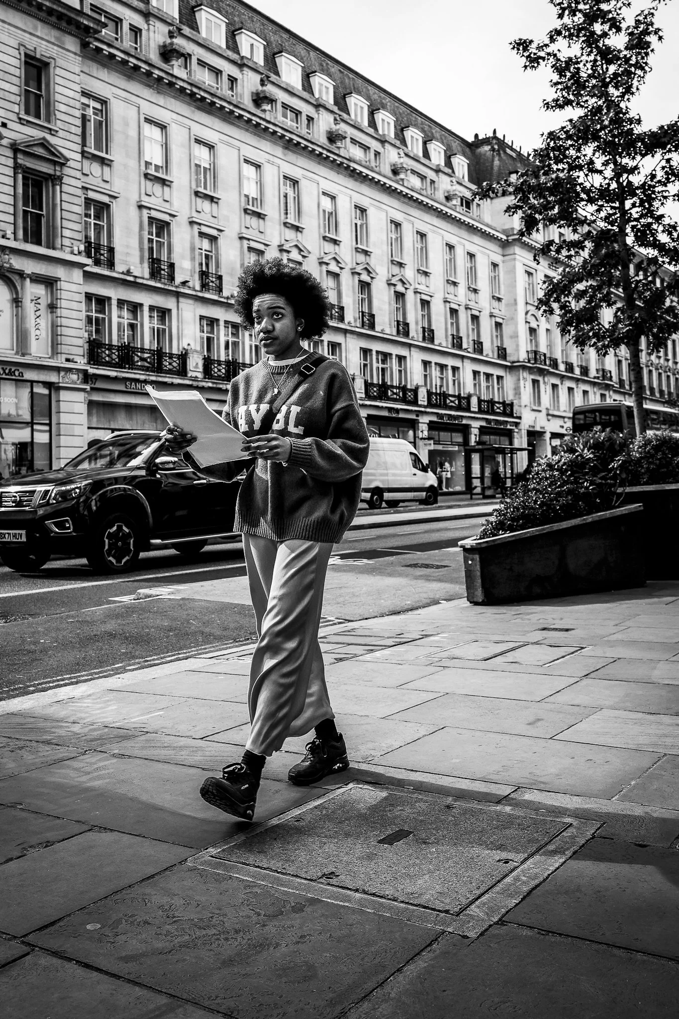 A Black woman with curly hair walking on a city sidewalk, holding papers, wearing a sweater and pants, with buildings, cars, and trees in the background, in black and white.
