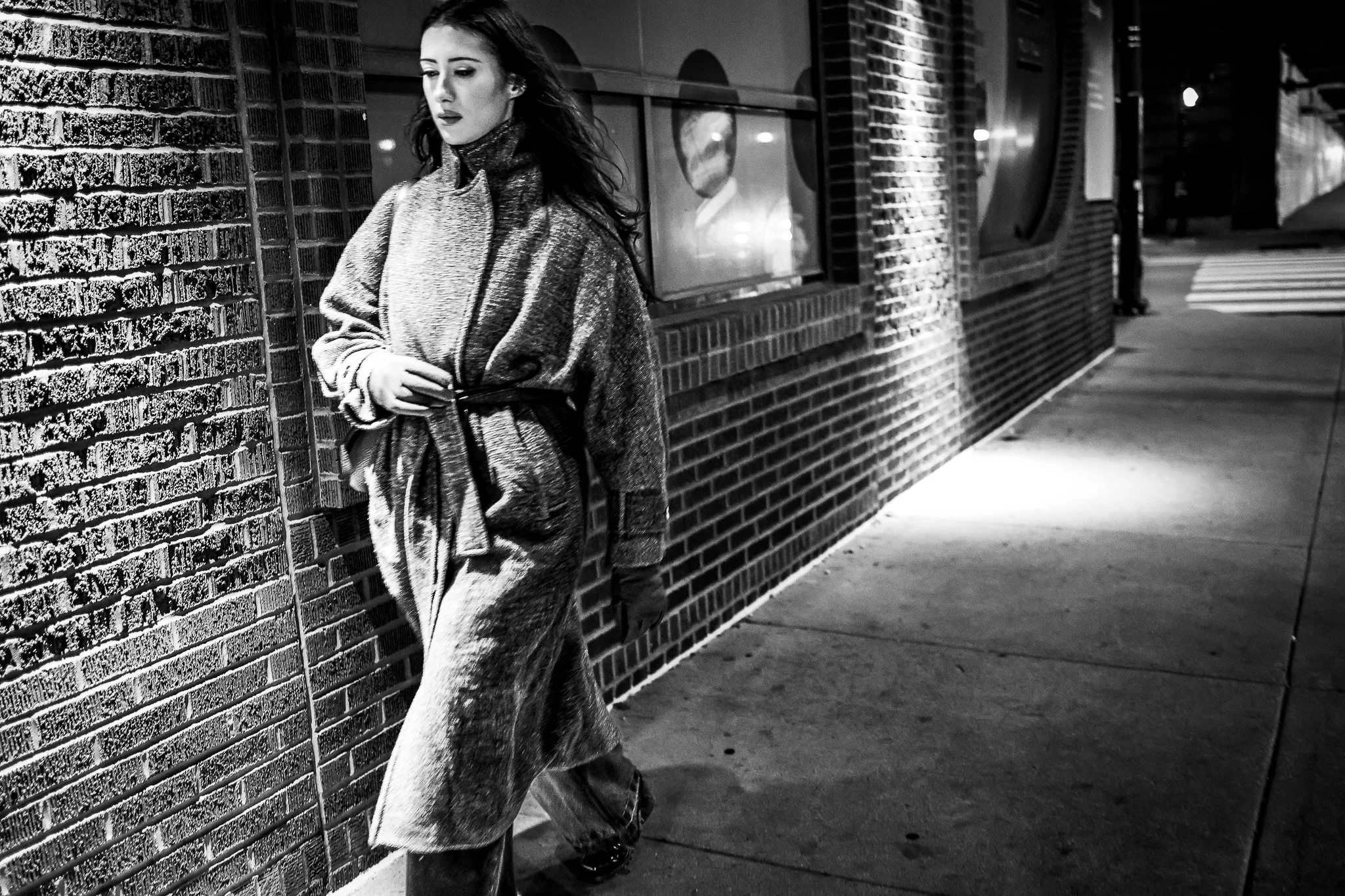 A woman with dark hair stands against a brick wall at night, looking downward with a serious expression. She is wearing a long coat and pants, with hands in her coat pockets. The scene is in black and white, with streetlights illuminating the sidewal