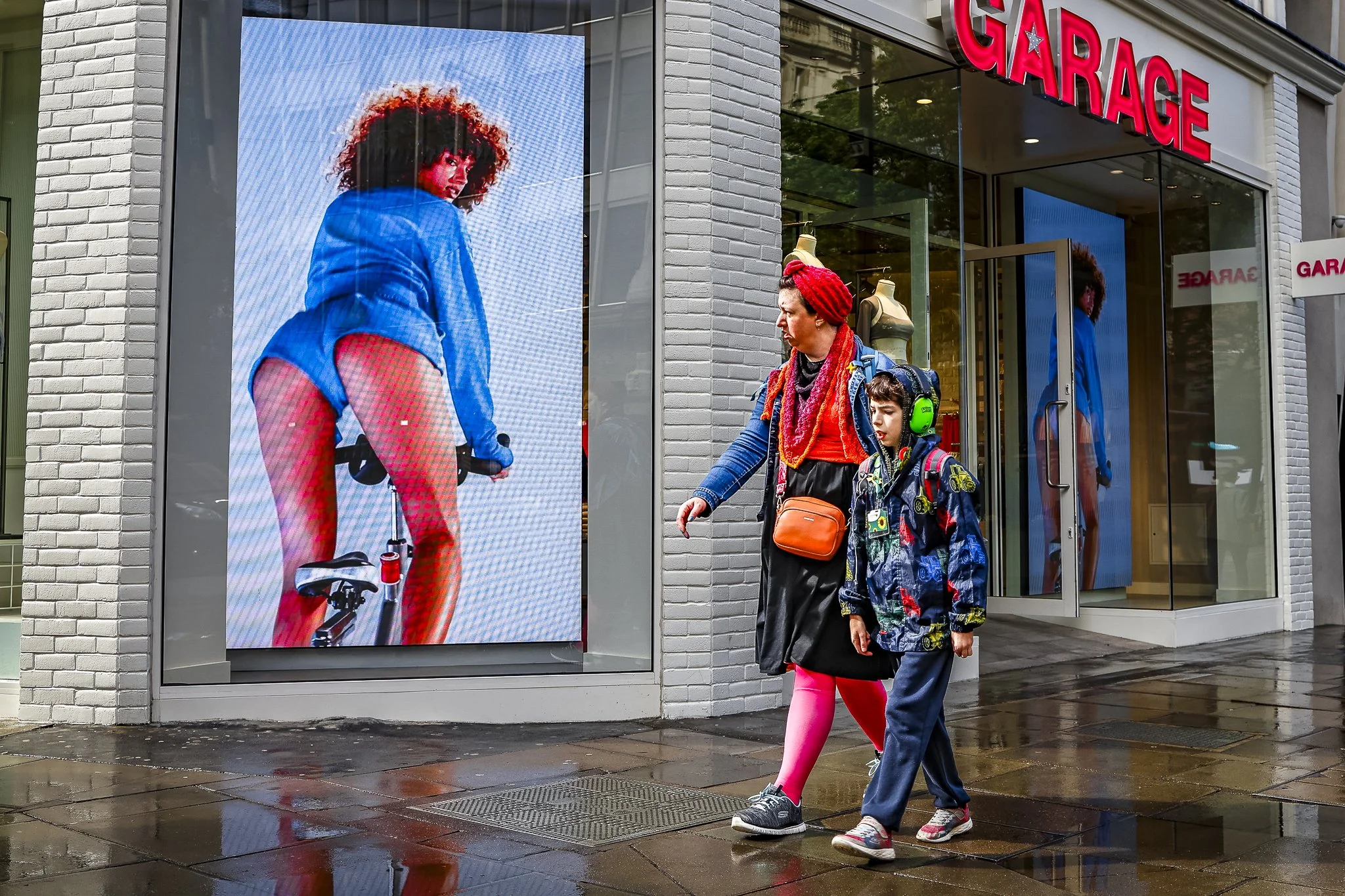 Two people walking past a storefront with a digital billboard showing a woman in a blue jacket and red pants, and the store has a sign that says "GARAGE" in large red letters.