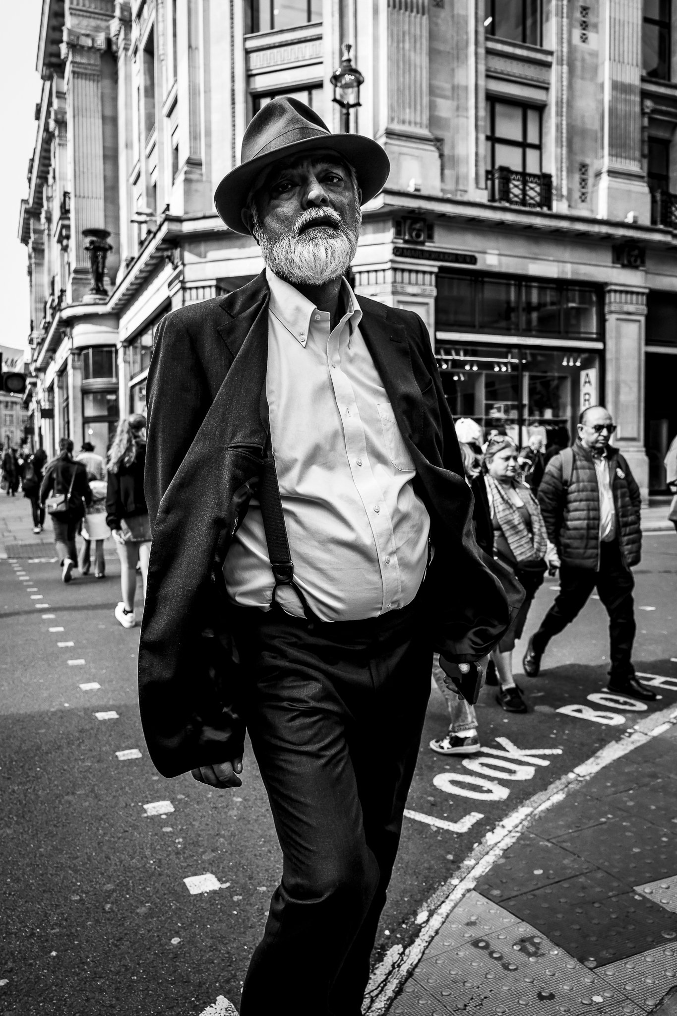 A black and white photo of an older man with a beard wearing a fedora hat, white shirt, and dark blazer, walking across a busy city street. Several people are in the background, and tall buildings with ornate architecture are visible.
