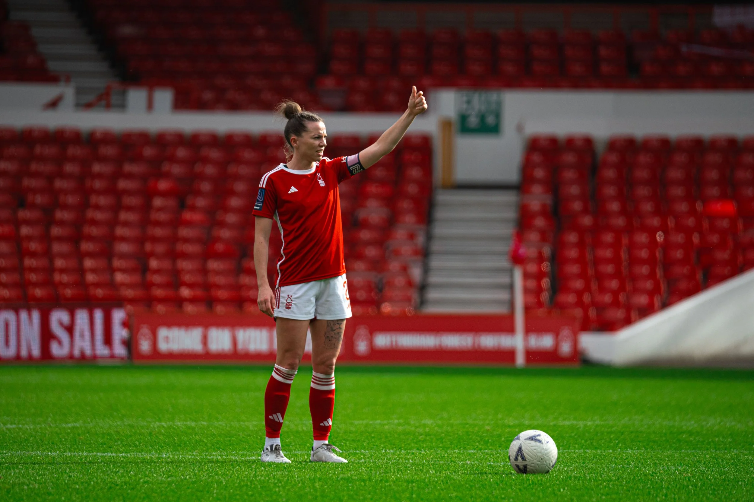 A female soccer player standing on the field preparing to kick a soccer ball, wearing a red jersey and shorts, with red socks, in a stadium with red seating.