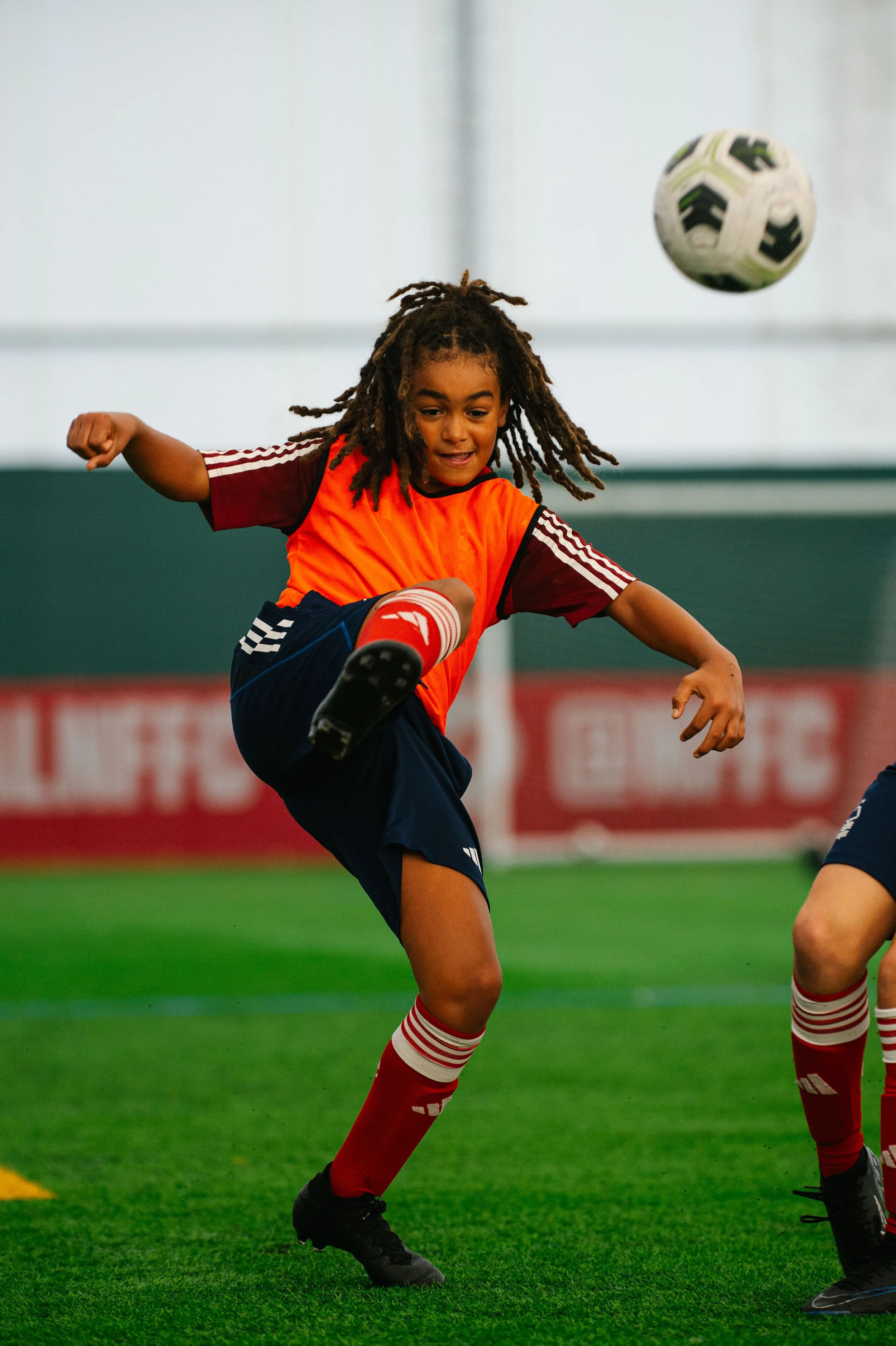 Child playing soccer on indoor field, kicking black and white ball in mid-air, wearing red and black sports uniform.