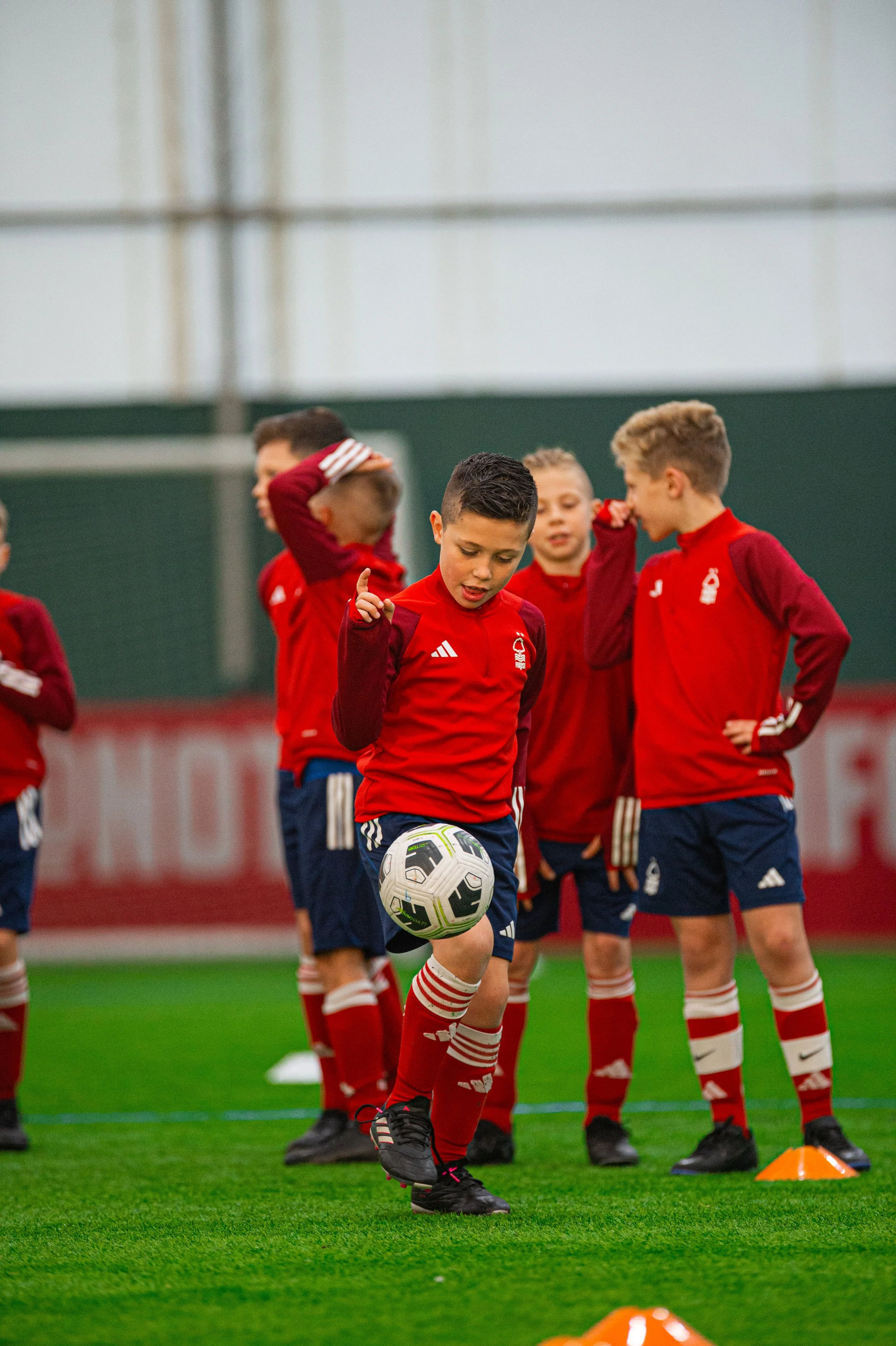 Children in red soccer uniforms practicing on an indoor field, with one boy controlling a soccer ball during a drill, and other kids watching.