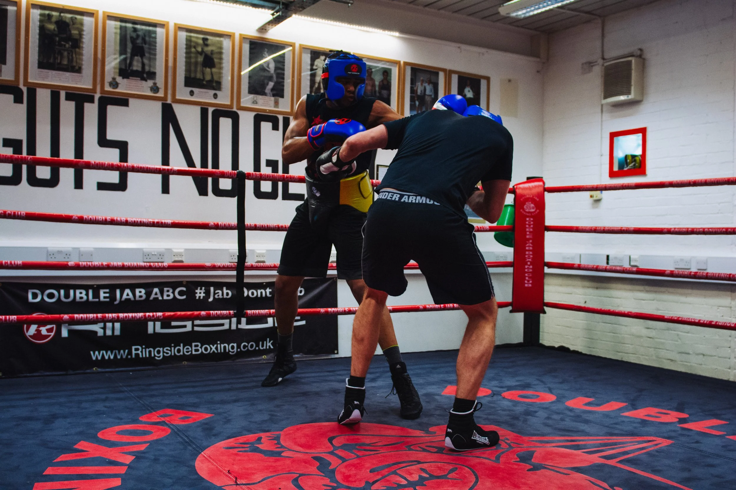 Two boxers training in a boxing gym, wearing protective gear and sparring inside a ring with red ropes, framed photos on the wall, and promotional banners on the ring floor.
