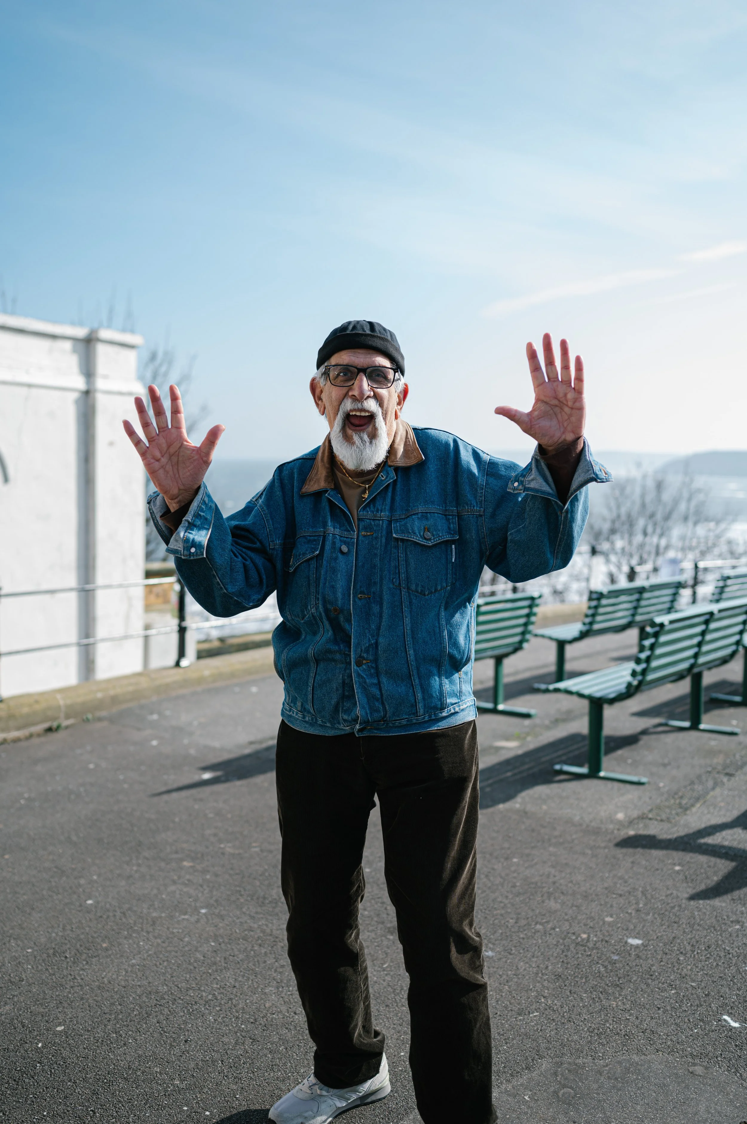 An elderly man with glasses, a black beanie, white beard, wearing a denim jacket, black pants, and white sneakers, standing outdoors on an overcast day with blue sky, benches, and a railing in the background, smiling and raising his hands.