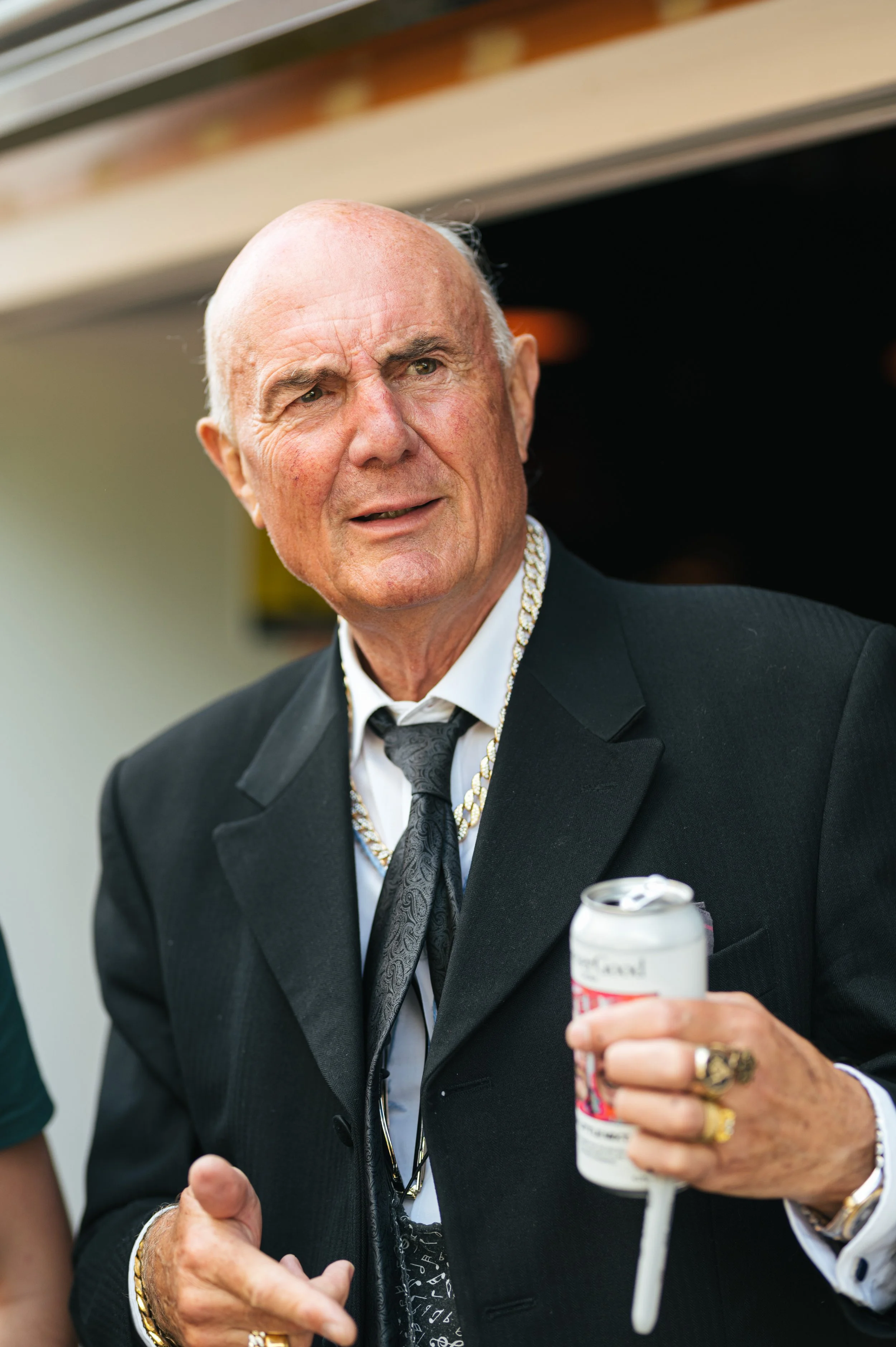 An elderly man wearing a black tuxedo with a silver patterned tie, gold jewelry, and holding a soda can with a straw. He appears to be at a social event or celebration.