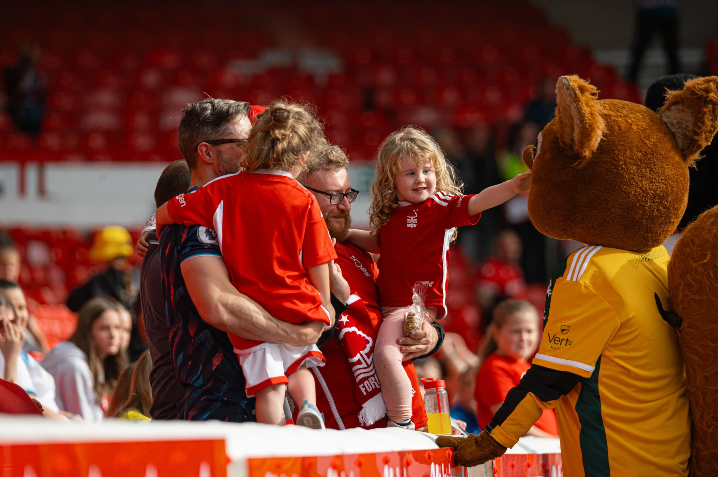 Children and adults, wearing red sports jerseys, interact with a lion mascot dressed in a yellow jersey at a sports event with red stadium seats in the background.