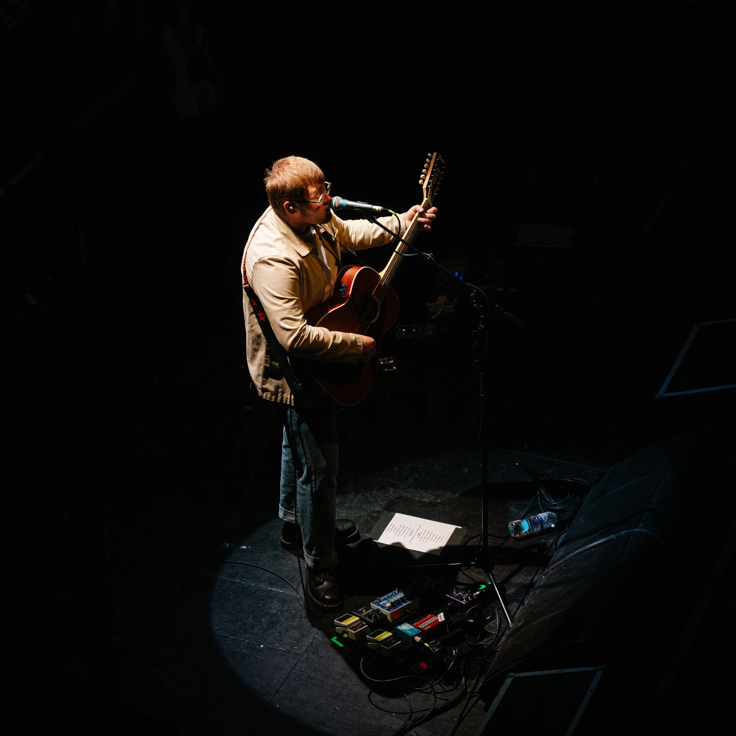 A musician performing on stage with an acoustic guitar, wearing glasses and headphones, surrounded by guitar effects pedals on the floor, with a dark background and minimal lighting.