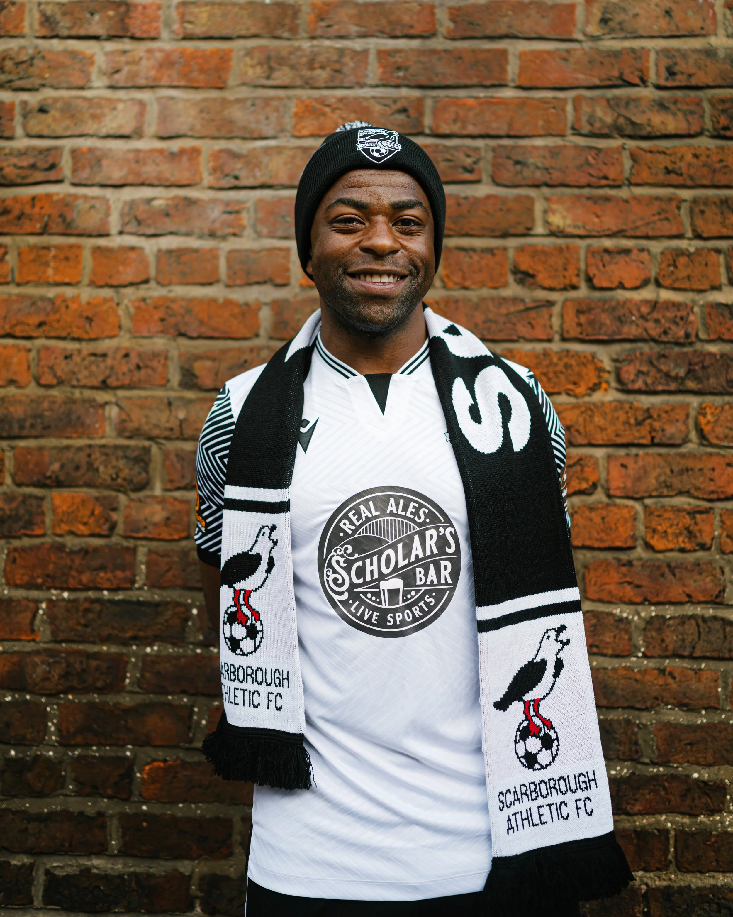 A man smiling in front of a brick wall, wearing a black beanie, a black and white scarf, a white sports jersey with a soccer ball and a circular logo, and black pants.