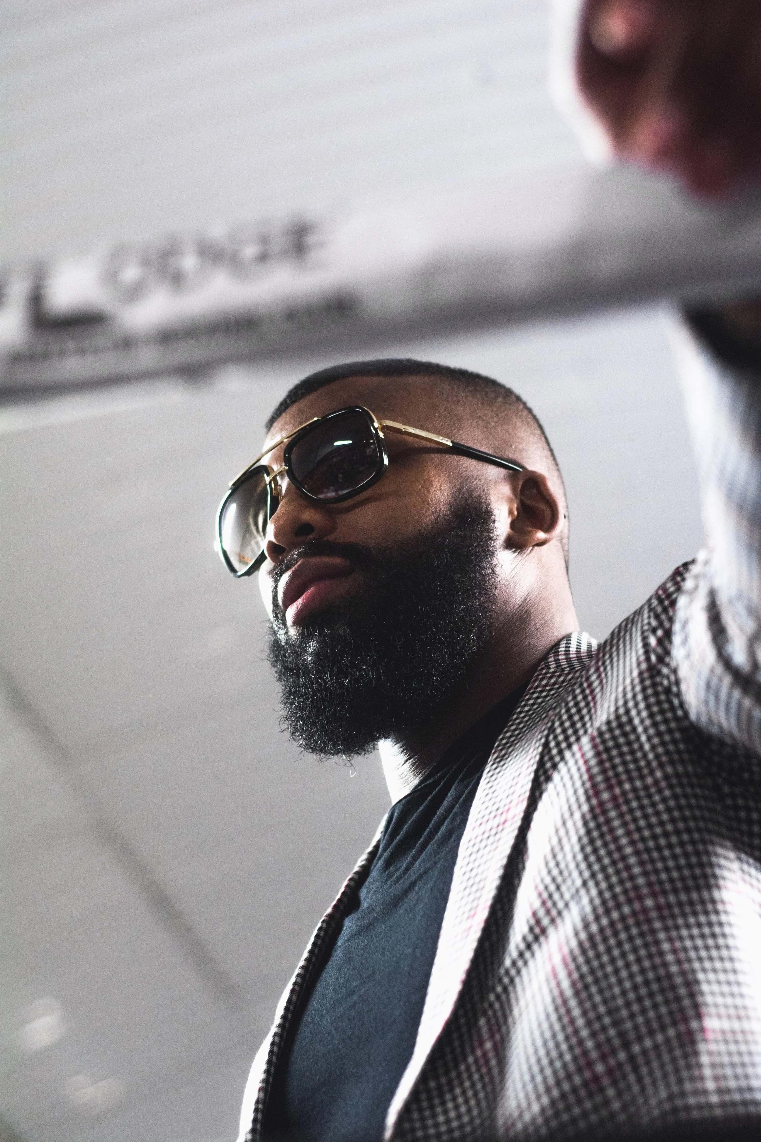 Close-up of a man with a beard wearing sunglasses and a checkered blazer, looking downward.
