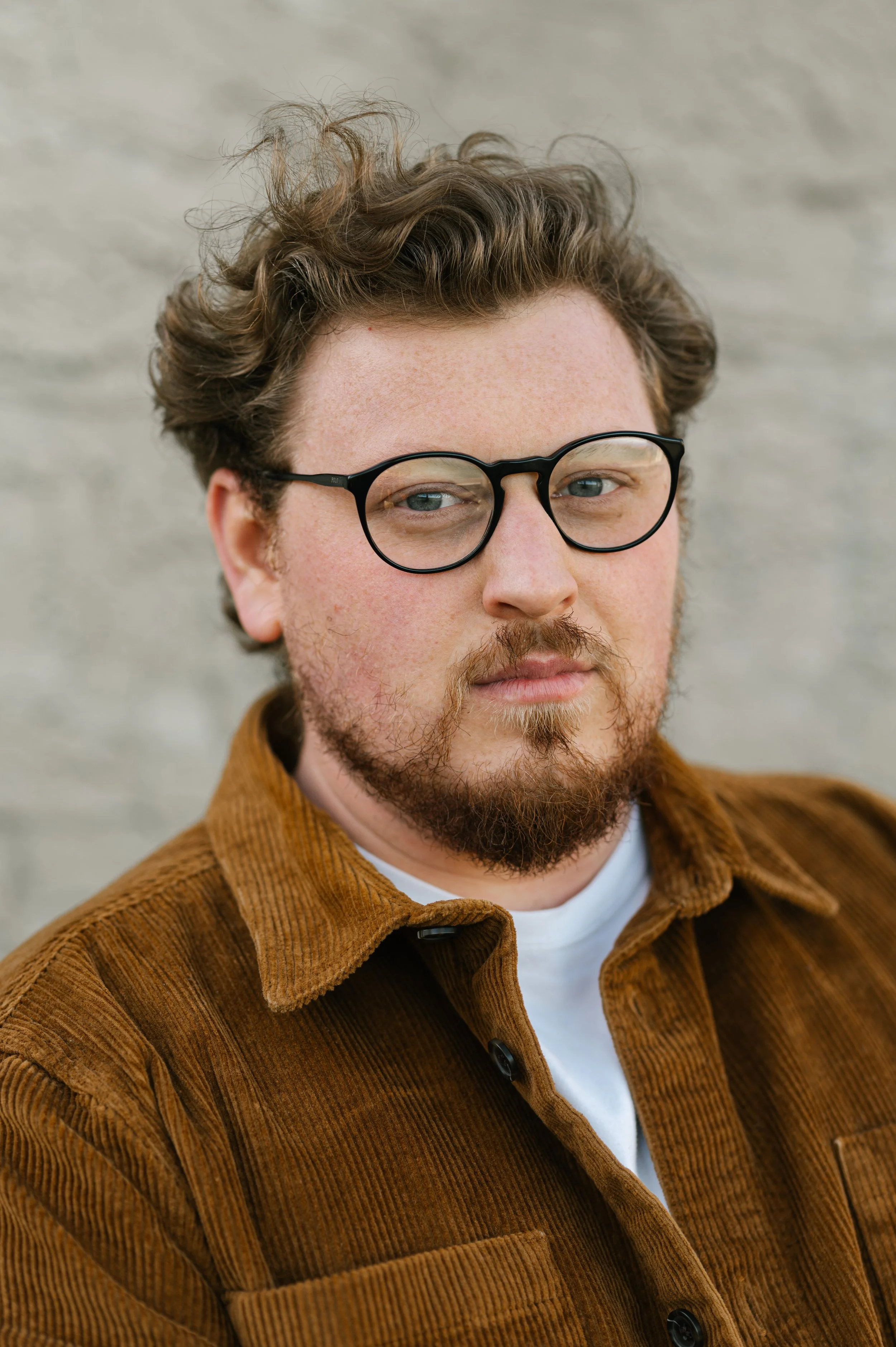 Close-up of a man with curly hair and a beard wearing black glasses, a white t-shirt, and a brown corduroy jacket, looking at the camera against a neutral background.