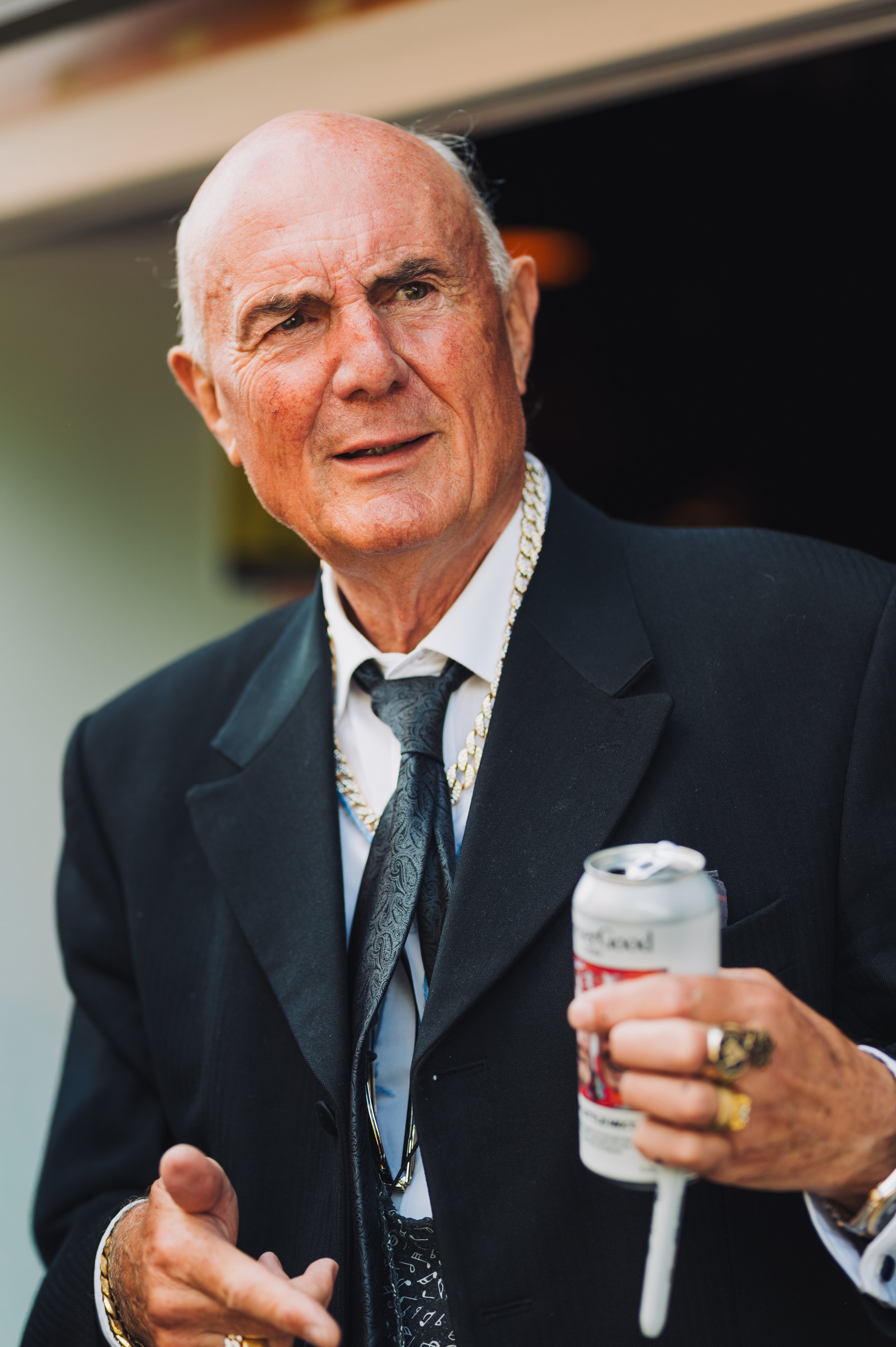 A middle-aged man with a bald head, dressed in a tuxedo, holding a can of beer, and wearing some jewelry, with lots of rings and bracelets. The man appears to be at an event or celebration.