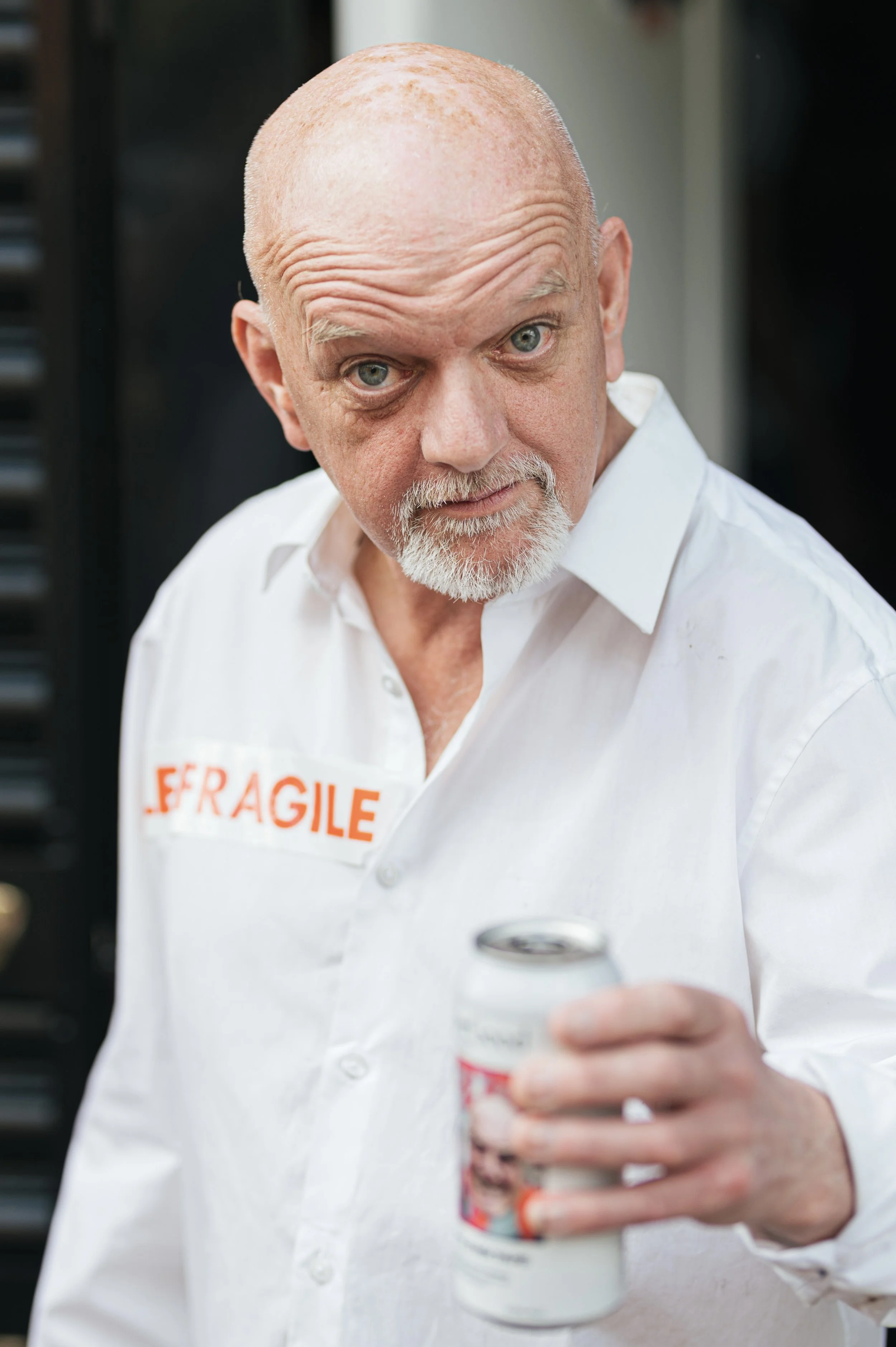 Older man with bald head and gray beard wearing a white shirt with a 'Fragile' sticker, holding a coffee cup, looking into the camera.