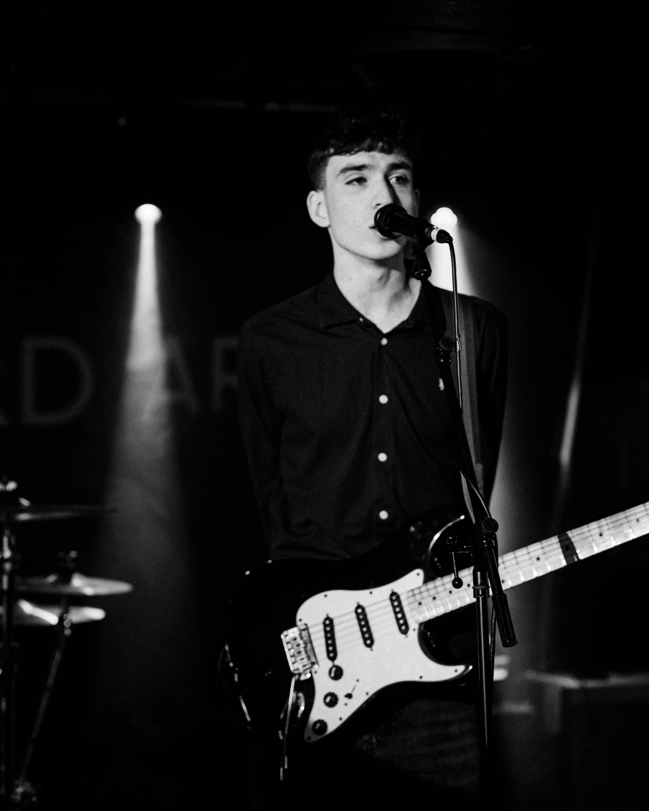 Black and white photo of a young male musician singing into a microphone while playing an electric guitar on stage with dark background.
