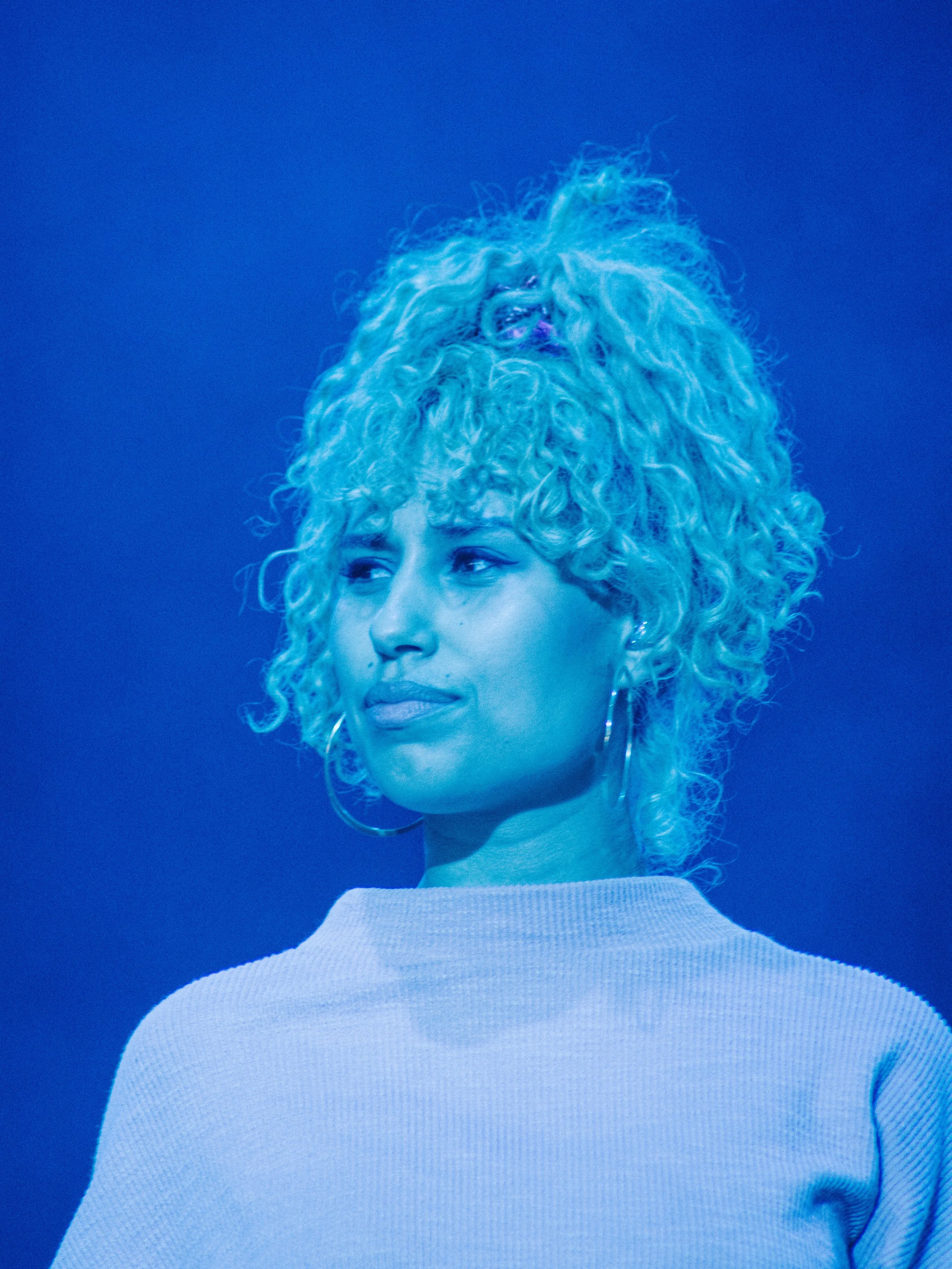 A woman with curly blonde hair, wearing a light-colored top and hoop earrings, looking to the side, against a plain blue background.
