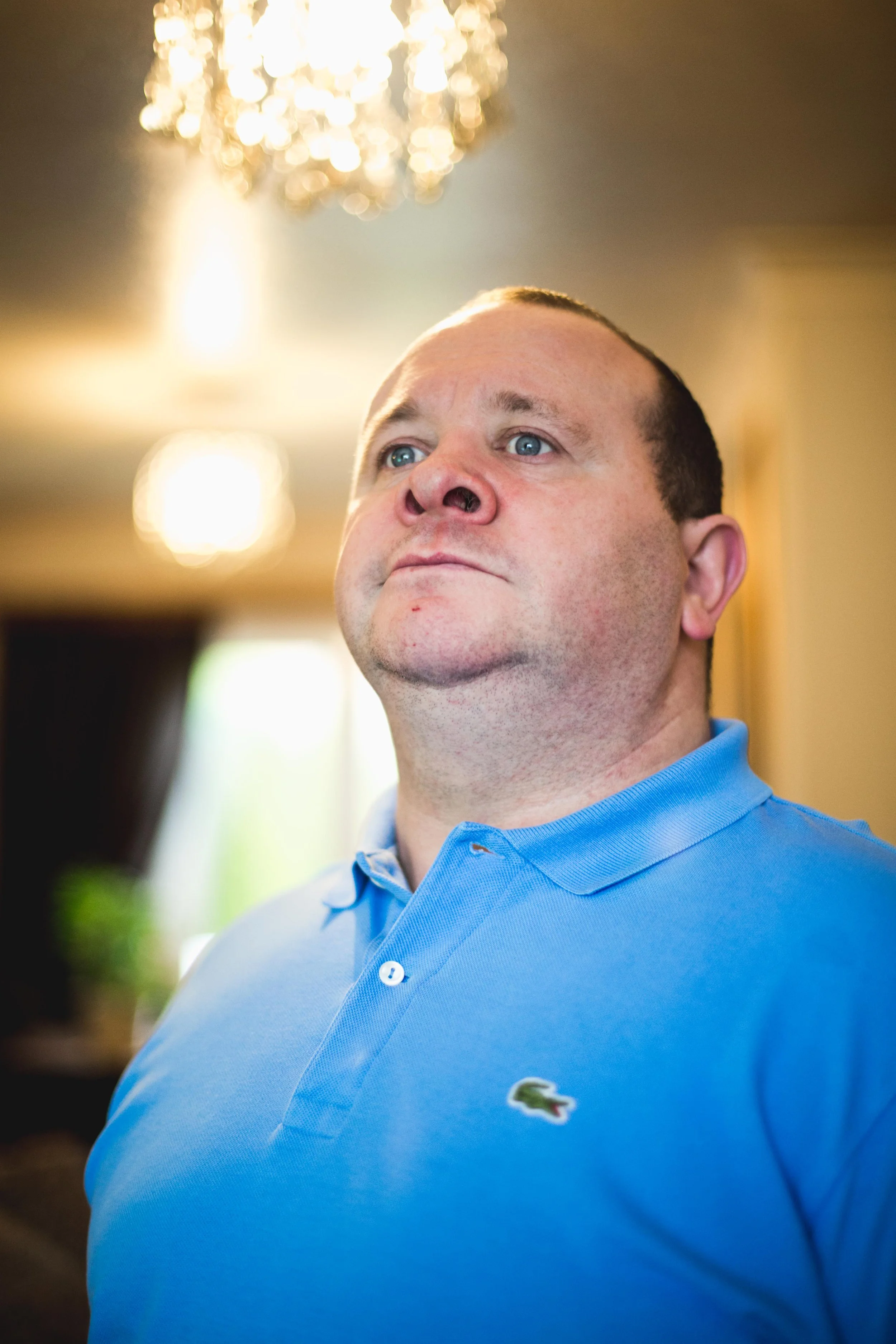A man with blue eyes and short hair wearing a light blue polo shirt with an embroidered crocodile logo. The background is out of focus with warm lighting and a chandelier hanging from the ceiling.