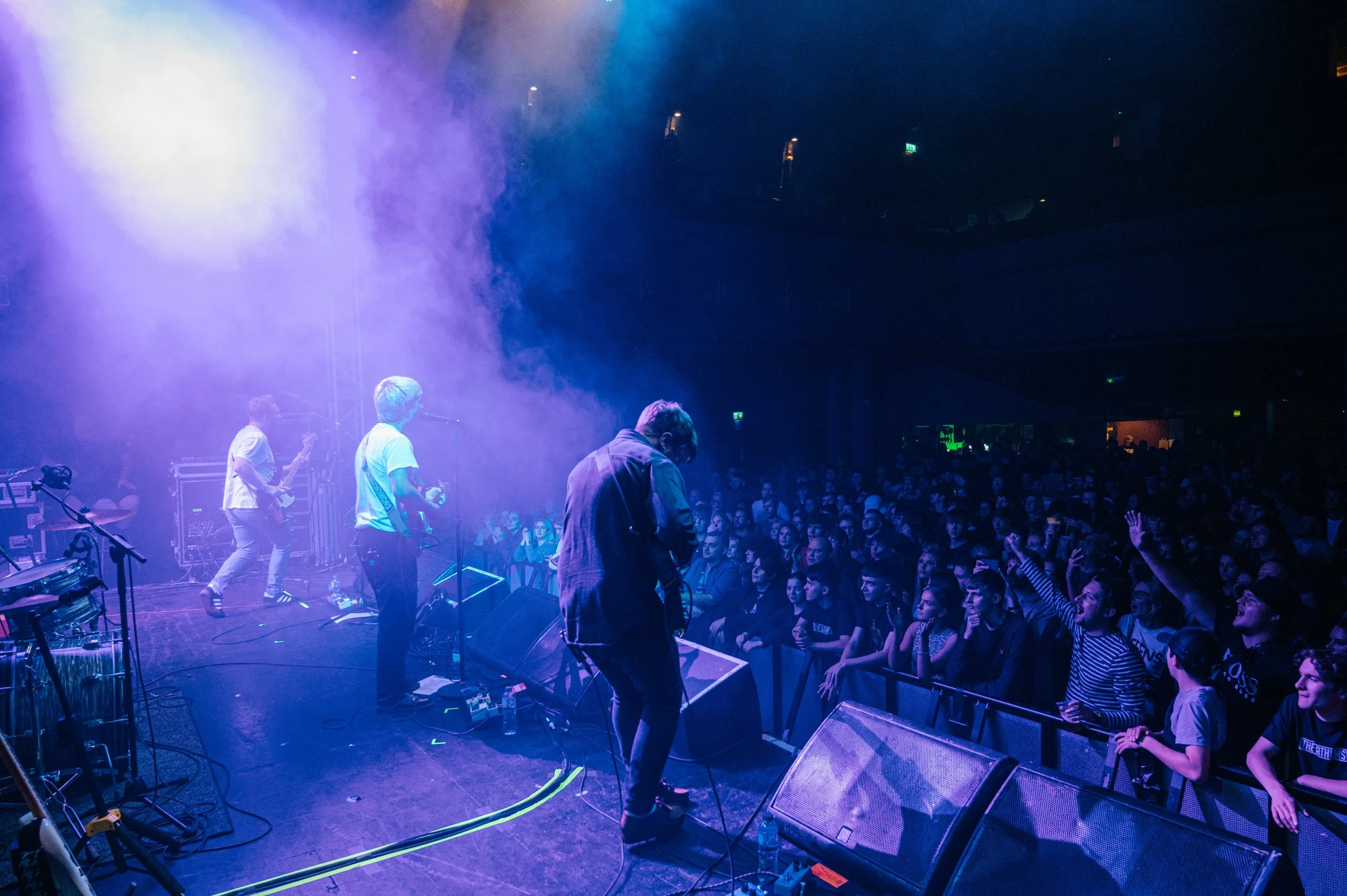 A band performing on stage with purple and blue lighting, in front of a large audience at a concert or music event.
