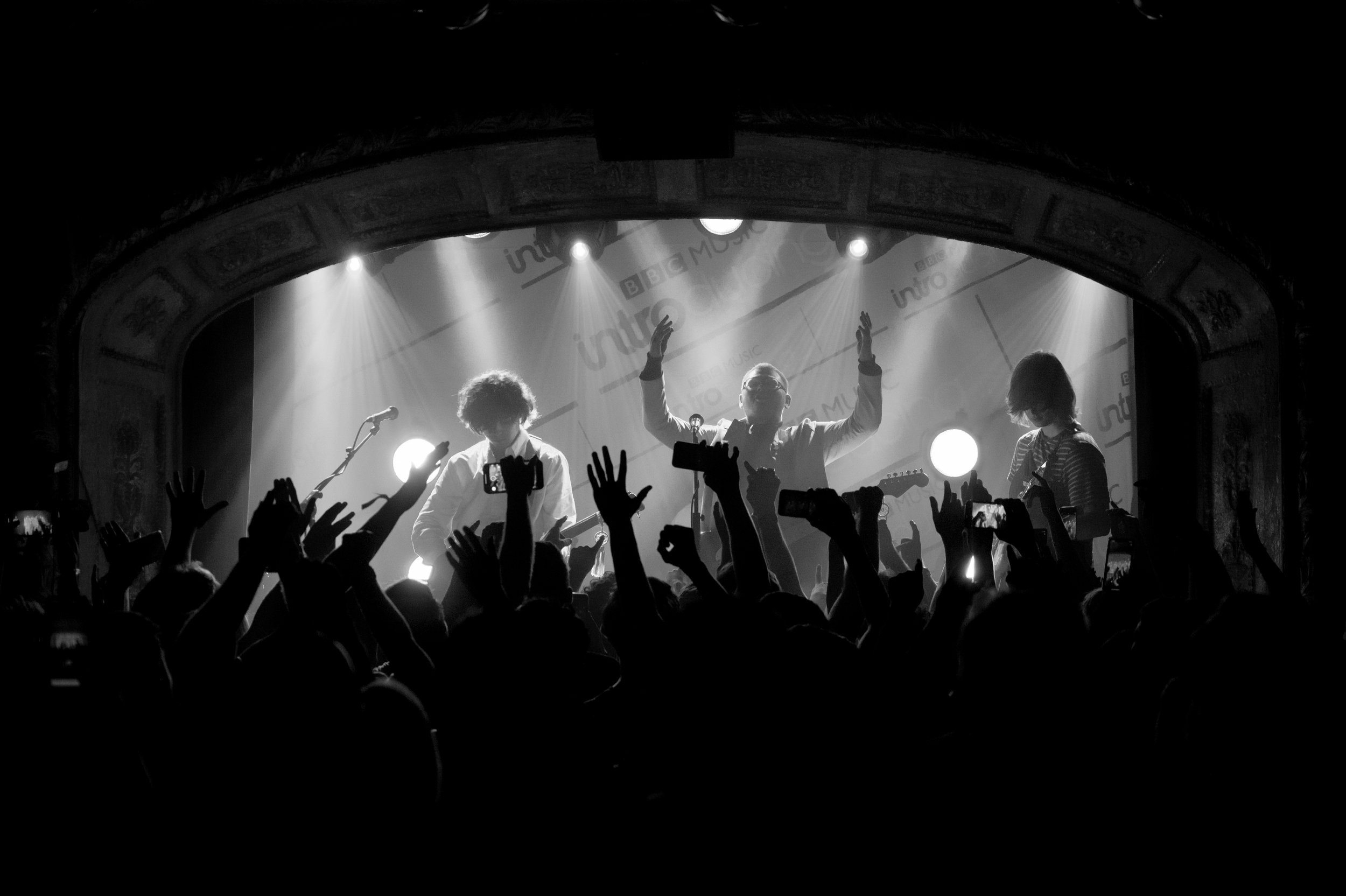 Black and white photo of a live music concert with a band playing on stage, audience raising hands, and stage lighting.