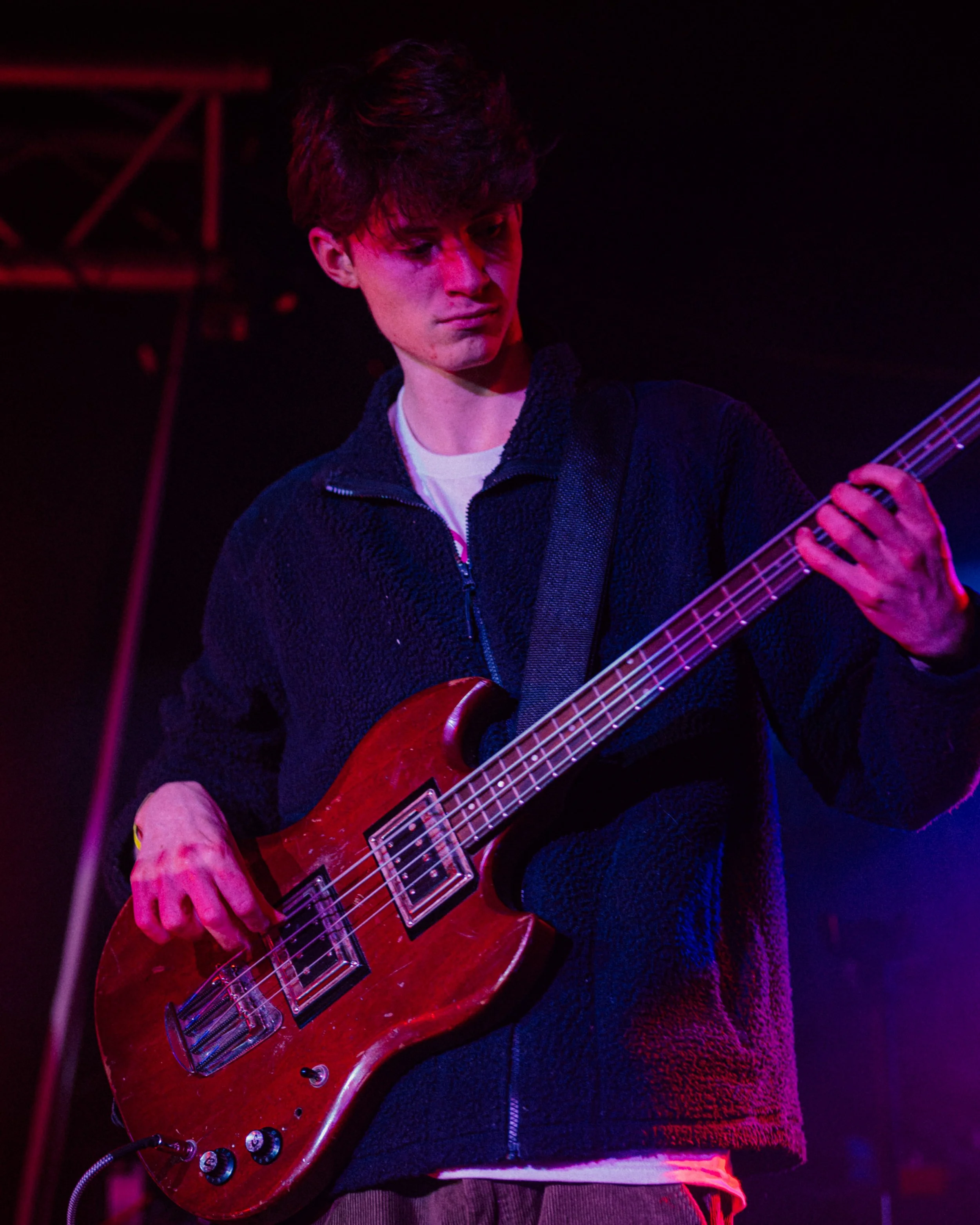 A young man playing an electric guitar on stage, illuminated by purple and red stage lights. He has dark hair, wears a black zip-up sweater, and holds the guitar with his left hand on the neck and his right hand near the body.