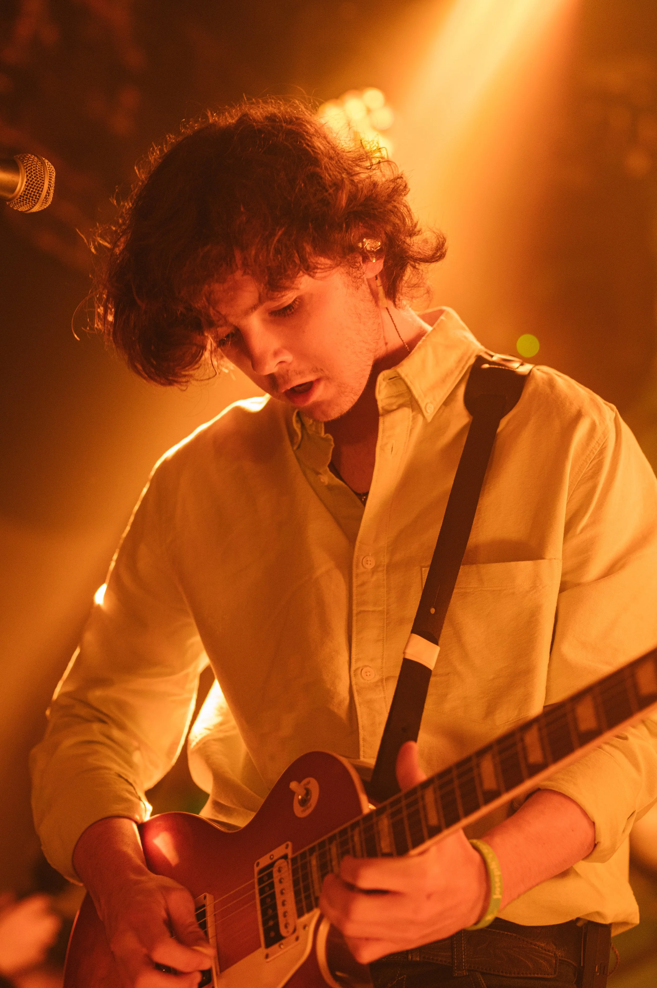A young man with curly dark hair playing an acoustic guitar, illuminated by warm orange stage lighting. He is wearing a light yellow shirt with rolled-up sleeves and a yellow bracelet.
