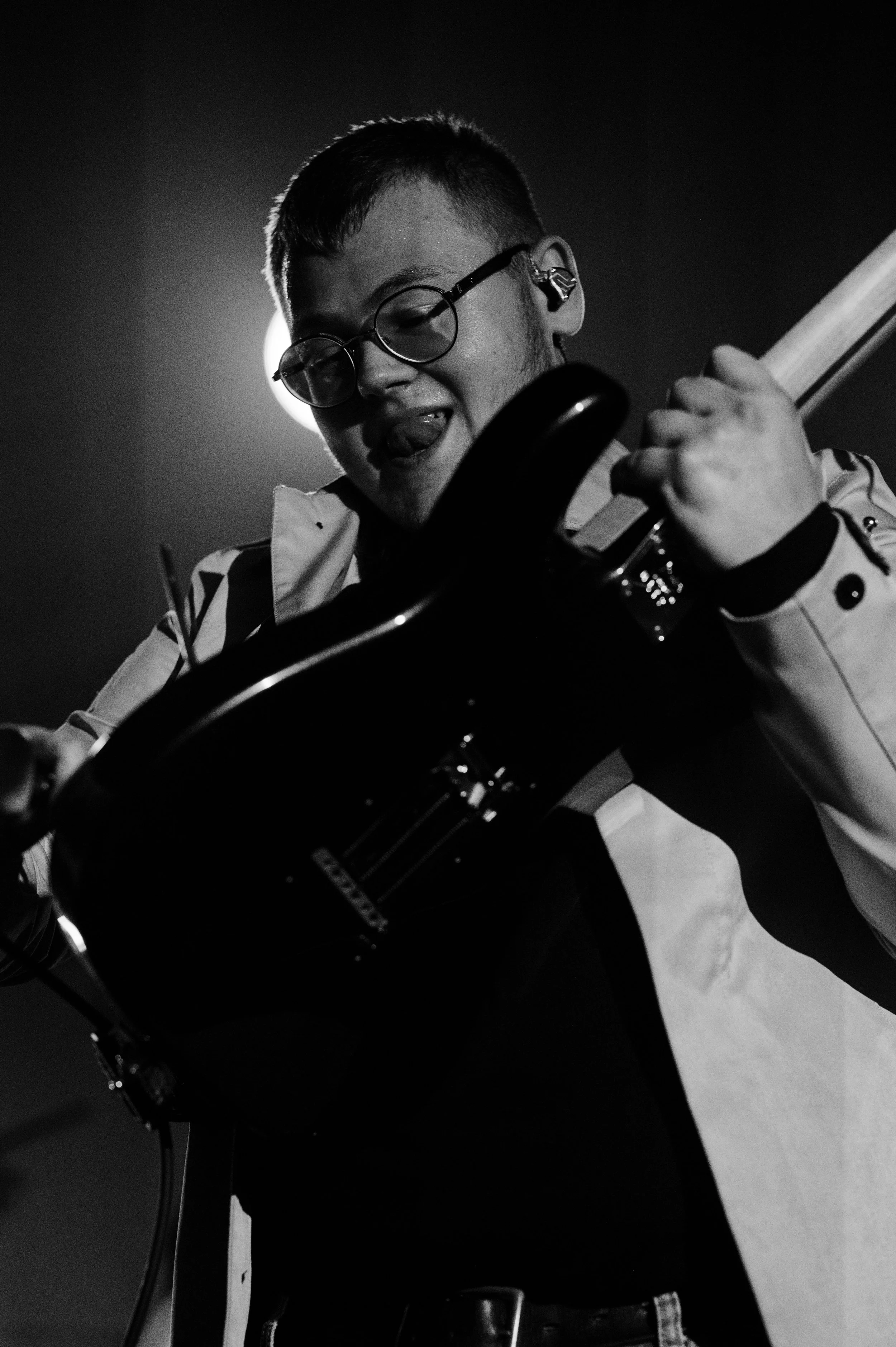 A person with short hair, glasses, and earrings is playing an electric guitar. The image is black and white with a bright light source behind the person's head.