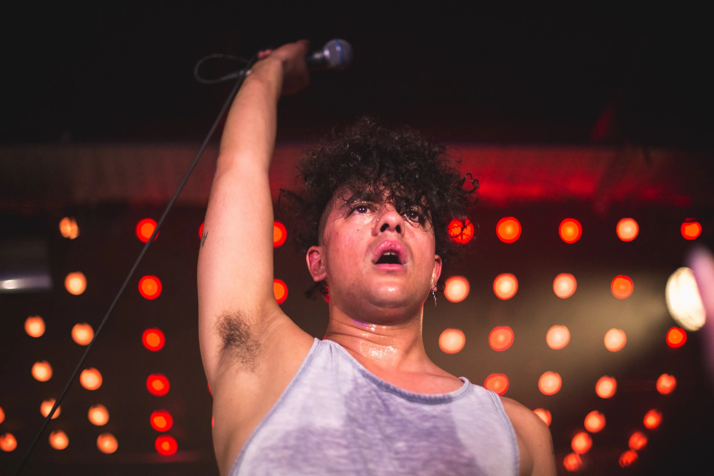 A person with curly hair and a sweat-drenched face is holding a microphone above their head on stage, with red stage lights in the background.