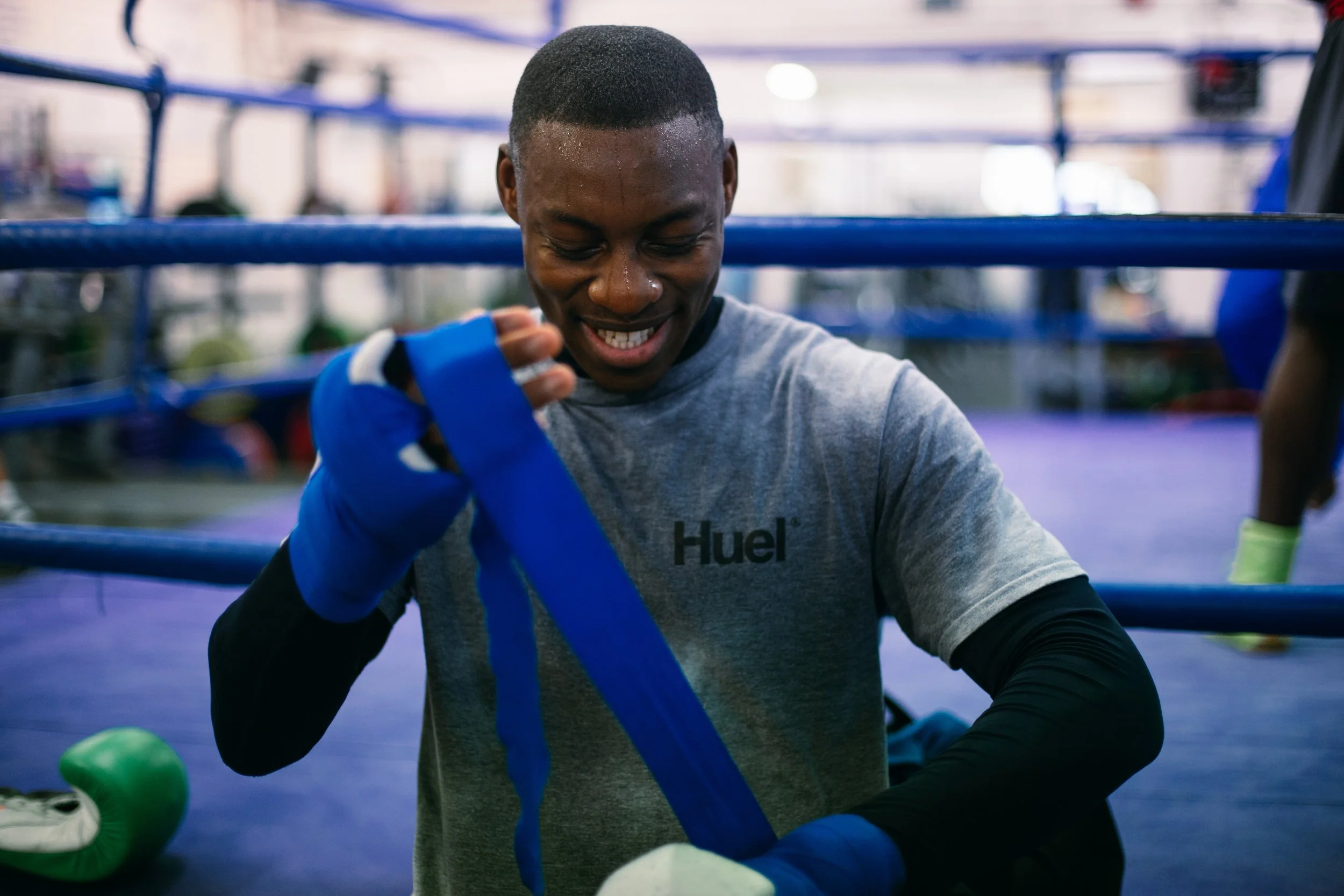 A male boxer with a close-cropped haircut, wearing a gray shirt with 'Huel' logo, wraps his hands with blue hand wraps inside a boxing gym.