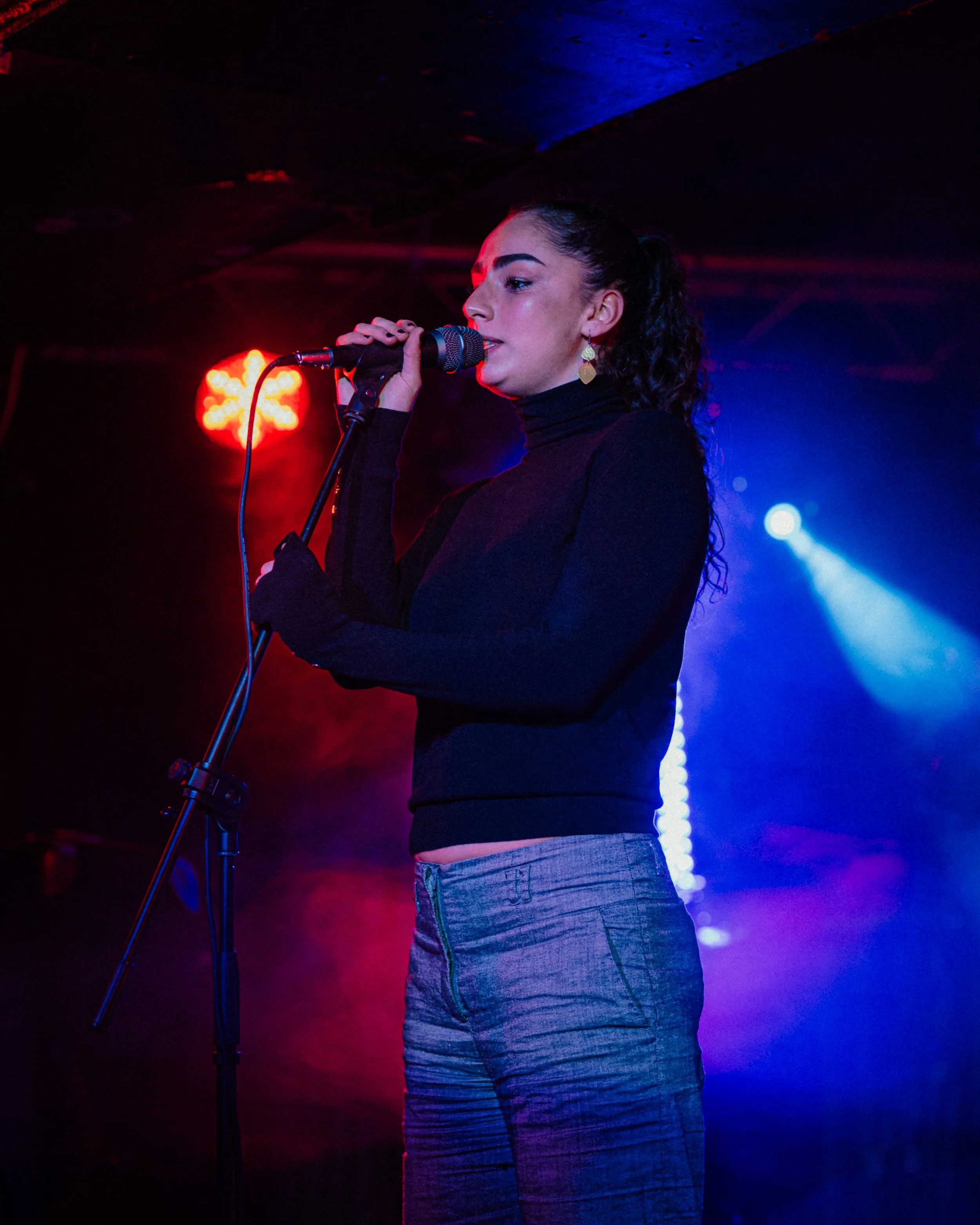A woman with curly dark hair, wearing a black turtleneck and jeans, is singing into a microphone on stage, with colorful stage lights shining behind her.