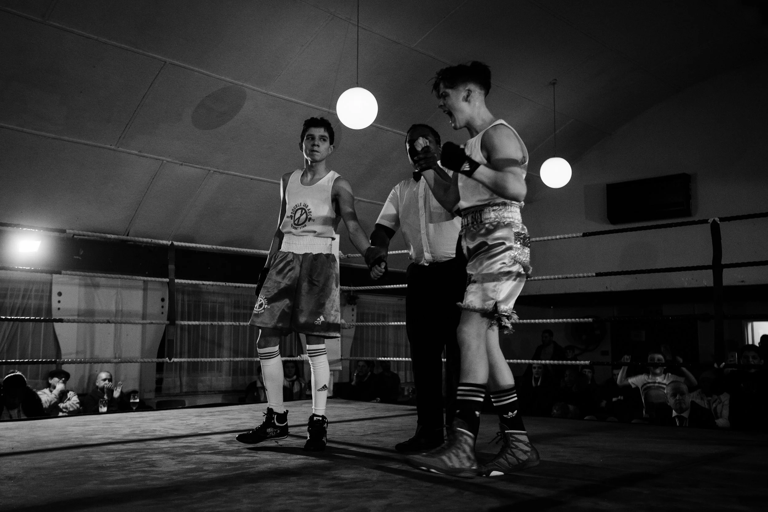 Black and white photo of two young boxers standing in the ring, with a referee between them. One boxer has his fists raised, while the other looks on, as the referee holds his hand. Spectators are sitting and standing around the ring in the backgroun