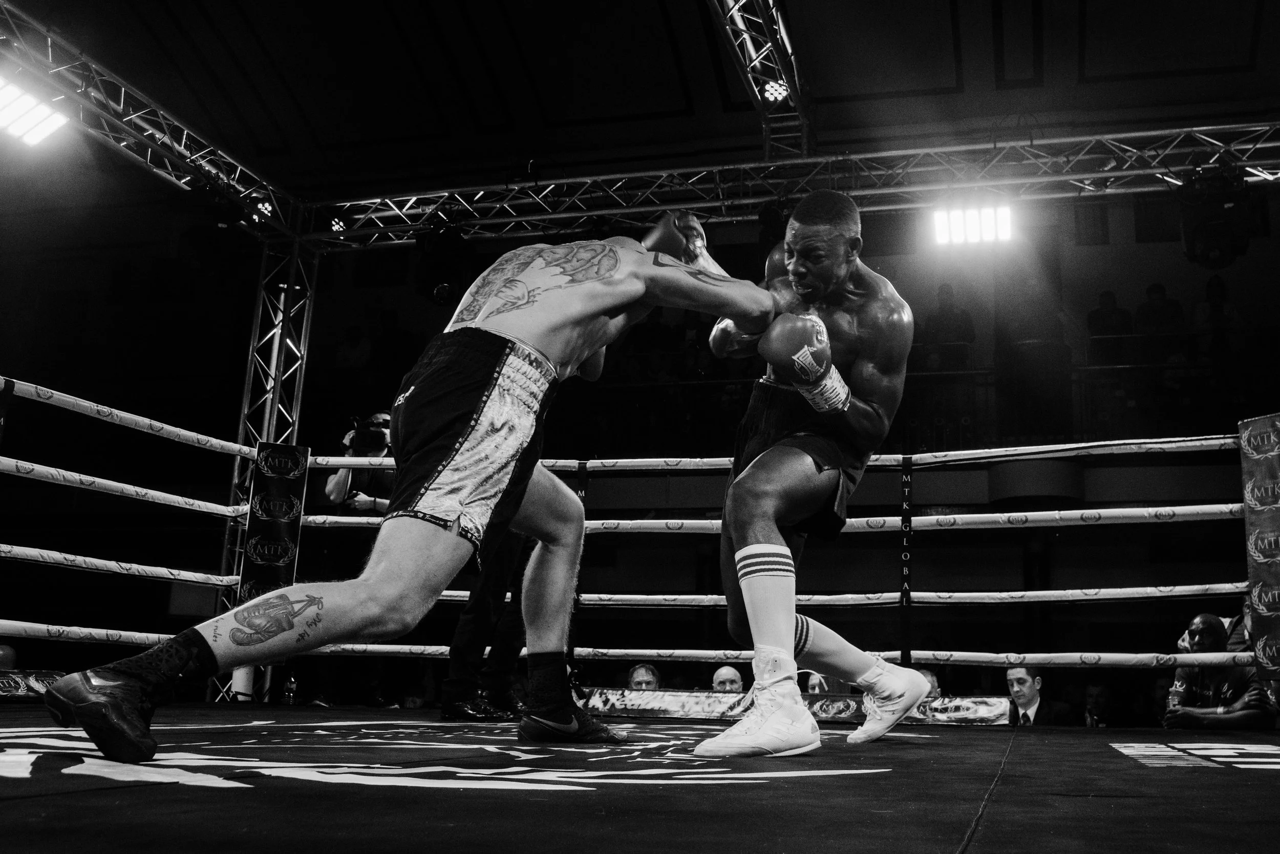 Black and white photo of two male boxers in a ring during a match, engaged in a punch fight with spectators watching.