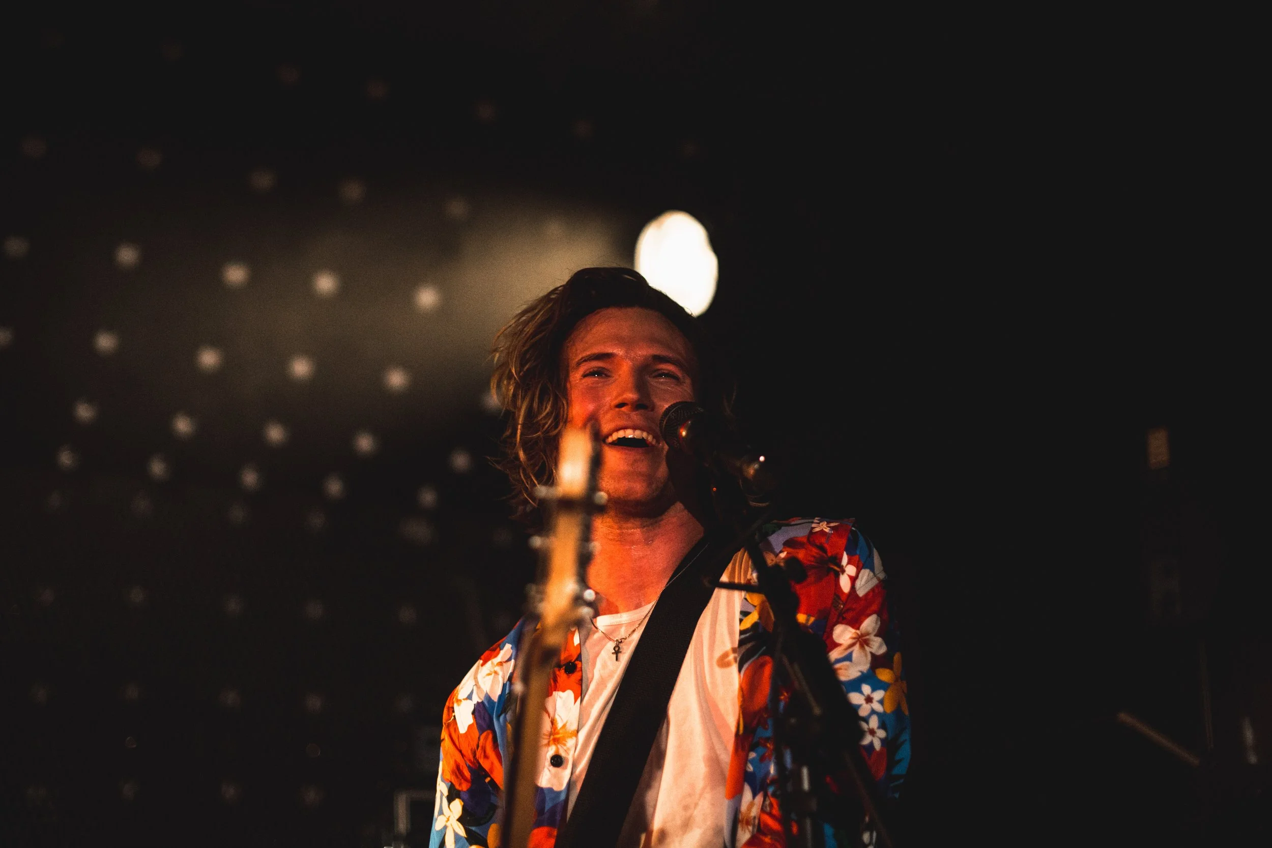 A person singing into a microphone on stage, wearing a colorful Hawaiian shirt, with stage lighting and a dark background.