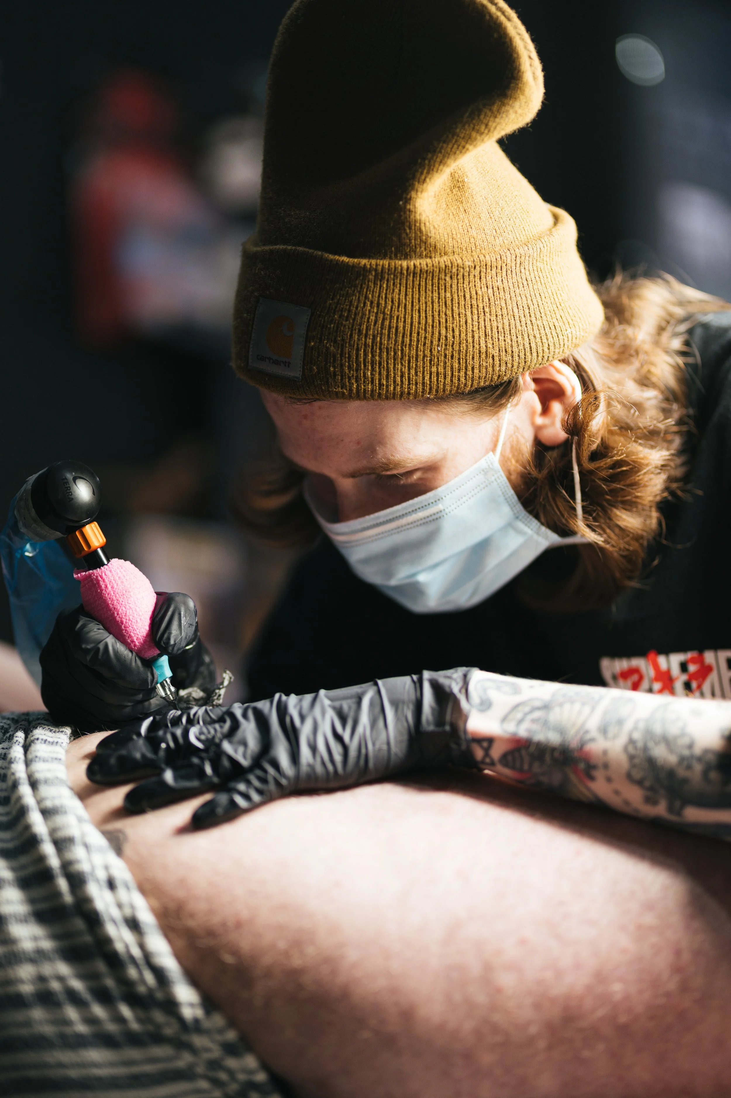 A person wearing a brown beanie, a face mask, and gloves tattooing on someone's arm in a tattoo studio.