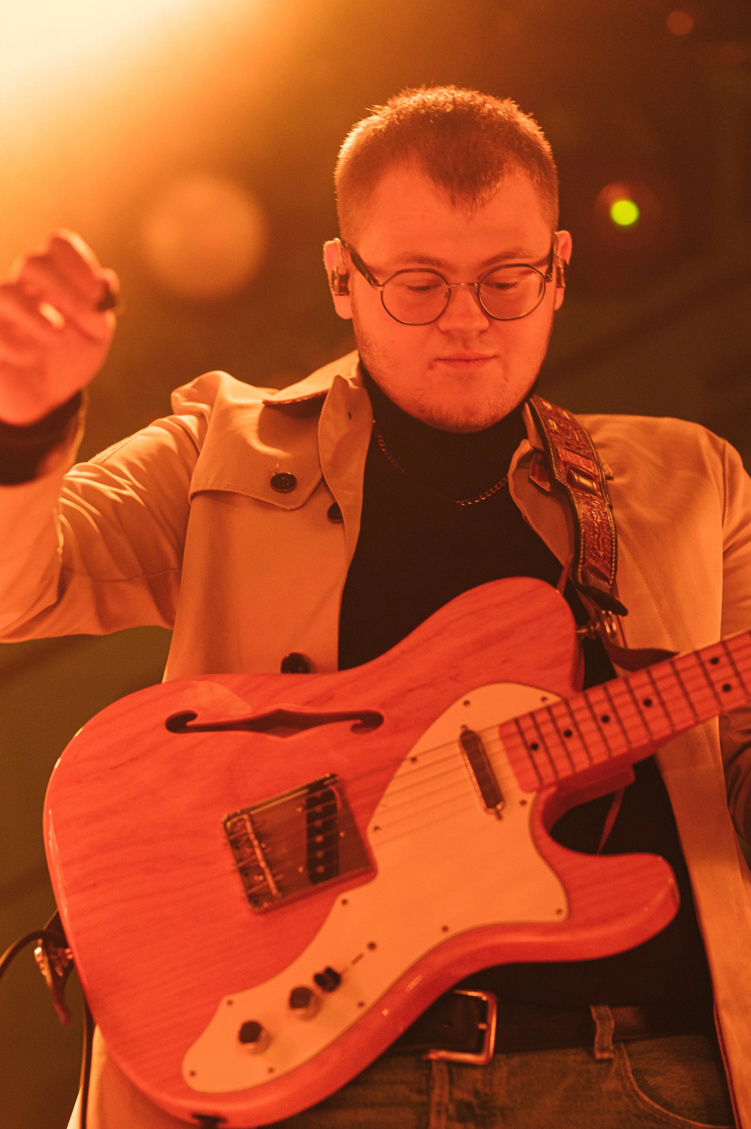 A person with short hair, glasses, and a beige jacket is playing an electric guitar in a warm, dimly lit environment.