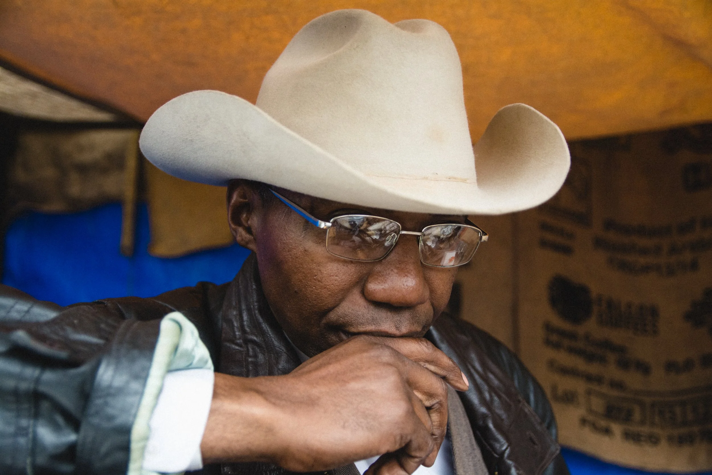 A man wearing a white cowboy hat and glasses, with a leather jacket, seated with his hand near his face, in a rustic setting.