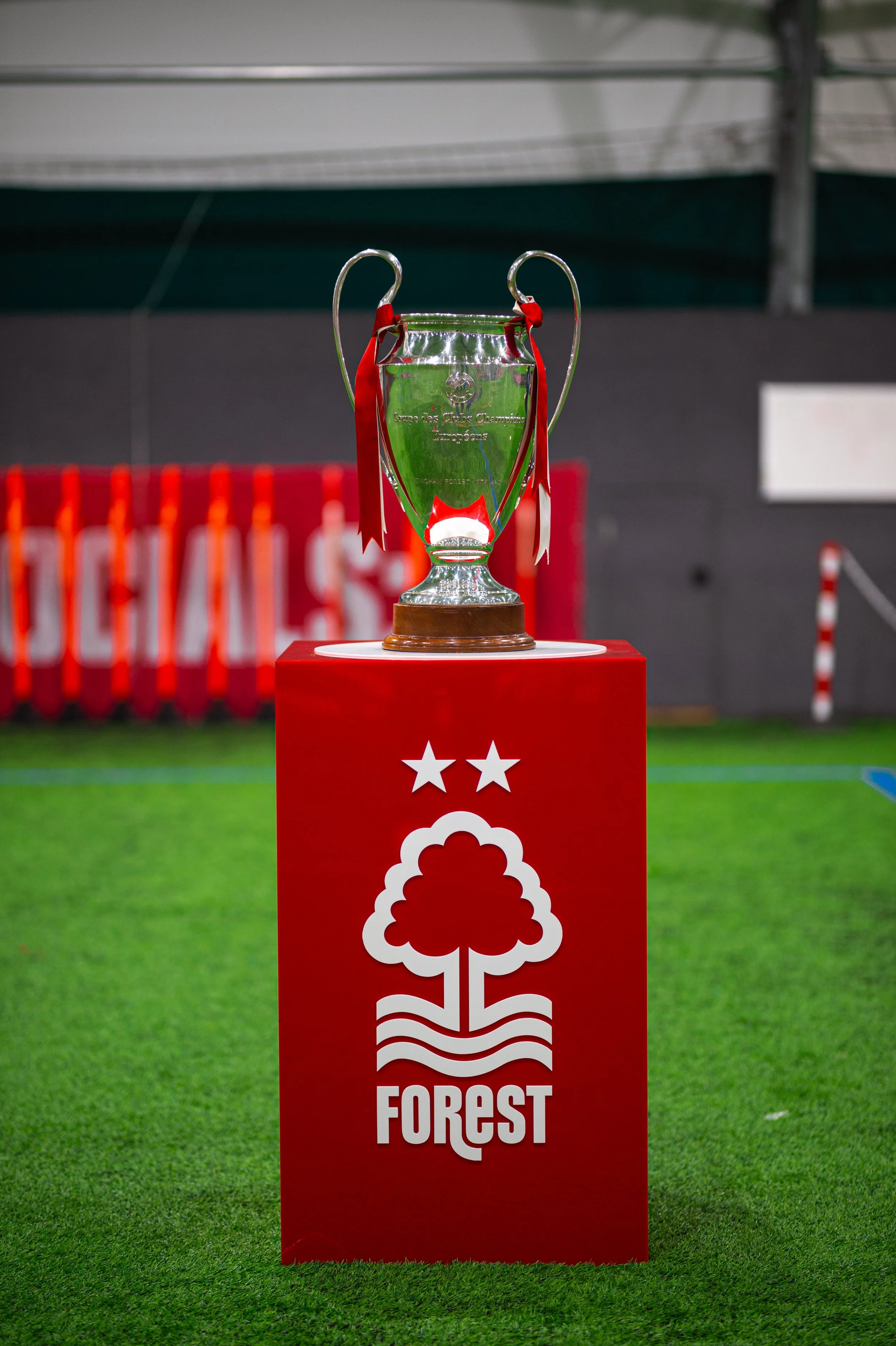 A silver trophy with red and white ribbons sits on a red pedestal covered with a cloth that has a white pine tree symbol and the word 'FORST' printed on it. The background shows a sports field and a sports hall.