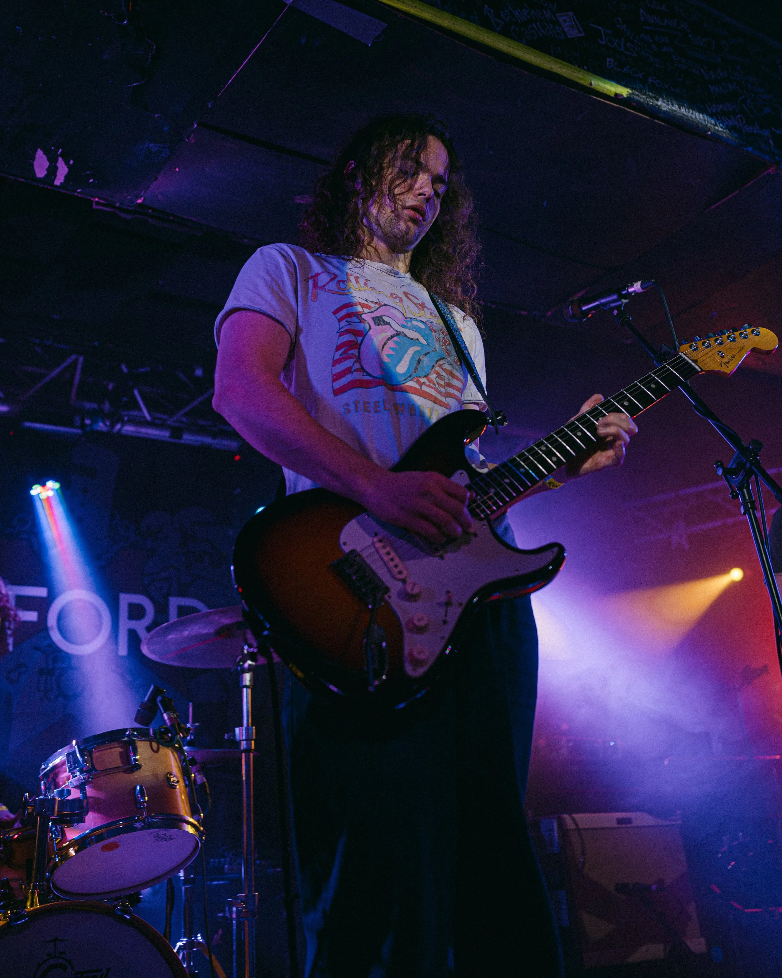 A person with long curly hair playing an electric guitar on stage, wearing a graphic T-shirt, with drums nearby and purple stage lighting.