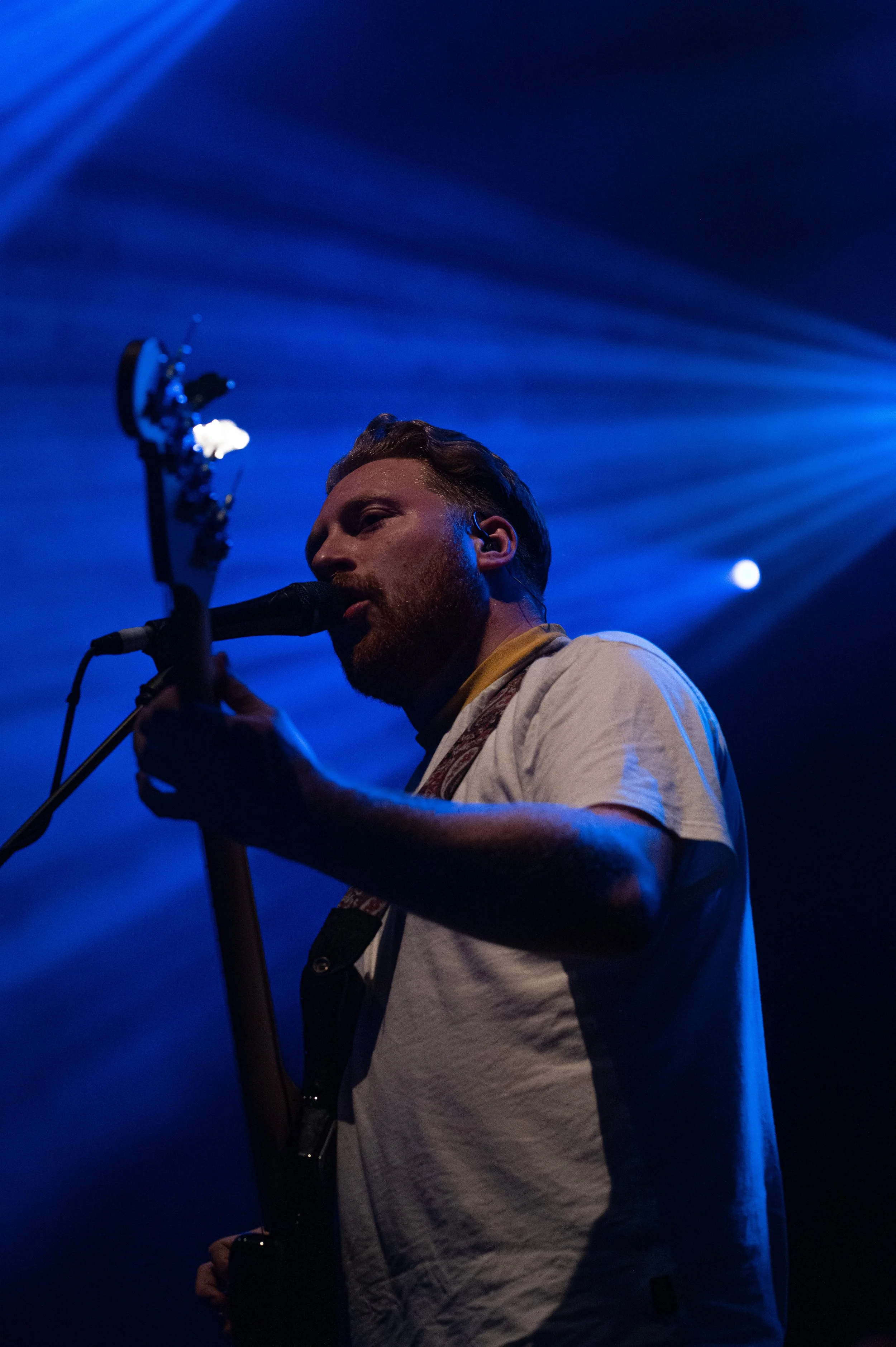 A man singing into a microphone with a guitar in his hand, performing on stage with blue light beams in the background.