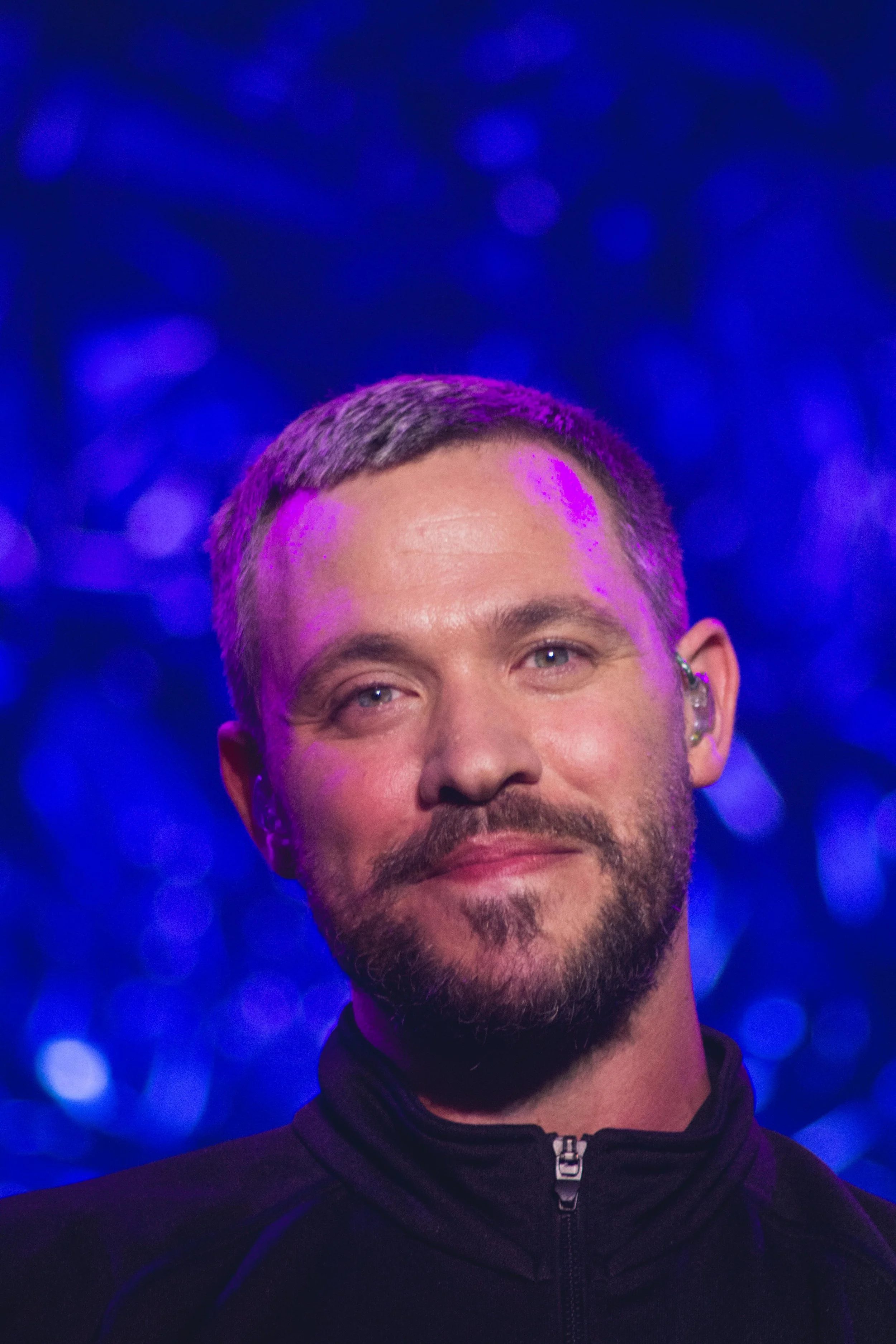 Close-up of a man with short hair, beard, and wearing a black zip-up jacket, smiling against a background of blue and purple bokeh lights.