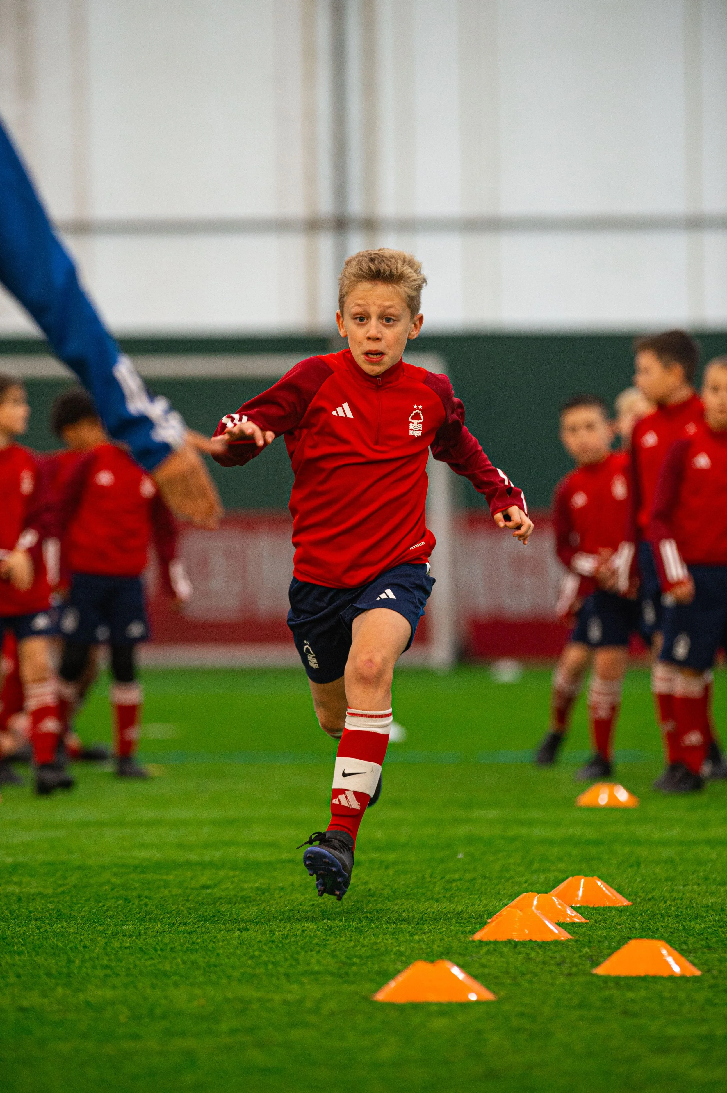 A young boy in a red soccer jersey and shorts is running on an indoor field, weaving through orange cone markers as part of a training drill, with soccer teammates in matching uniforms in the background.
