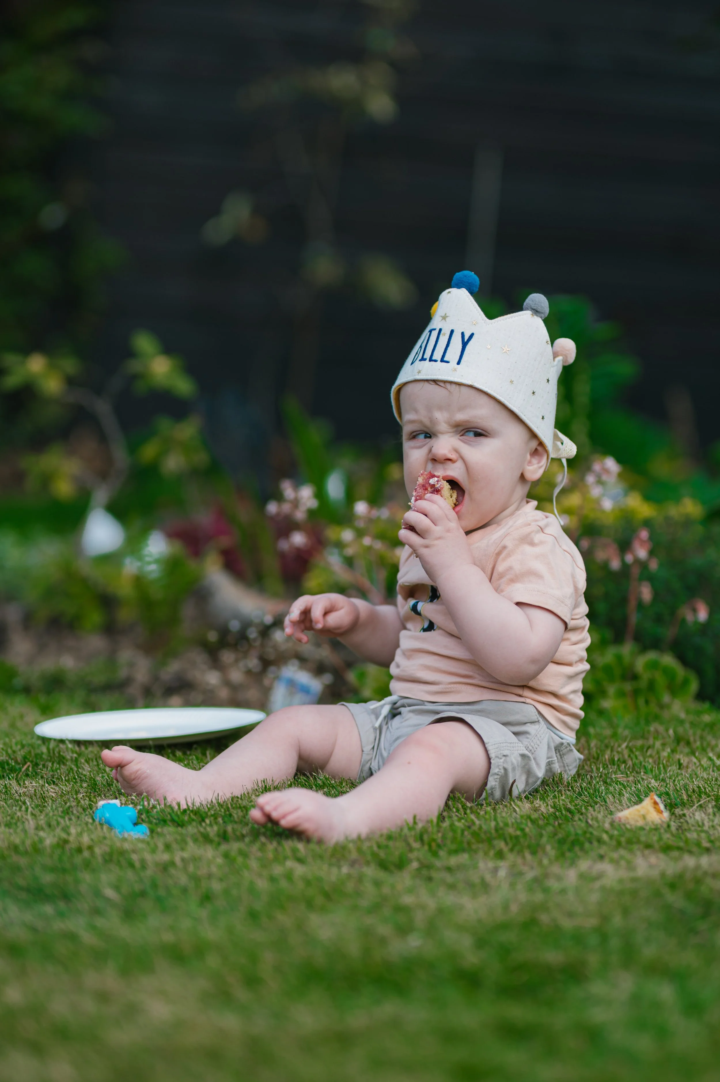 A young child sitting on grass outdoors, eating a piece of food, wearing a white hat with pom-poms and the name 'Billy' embroidered on it.