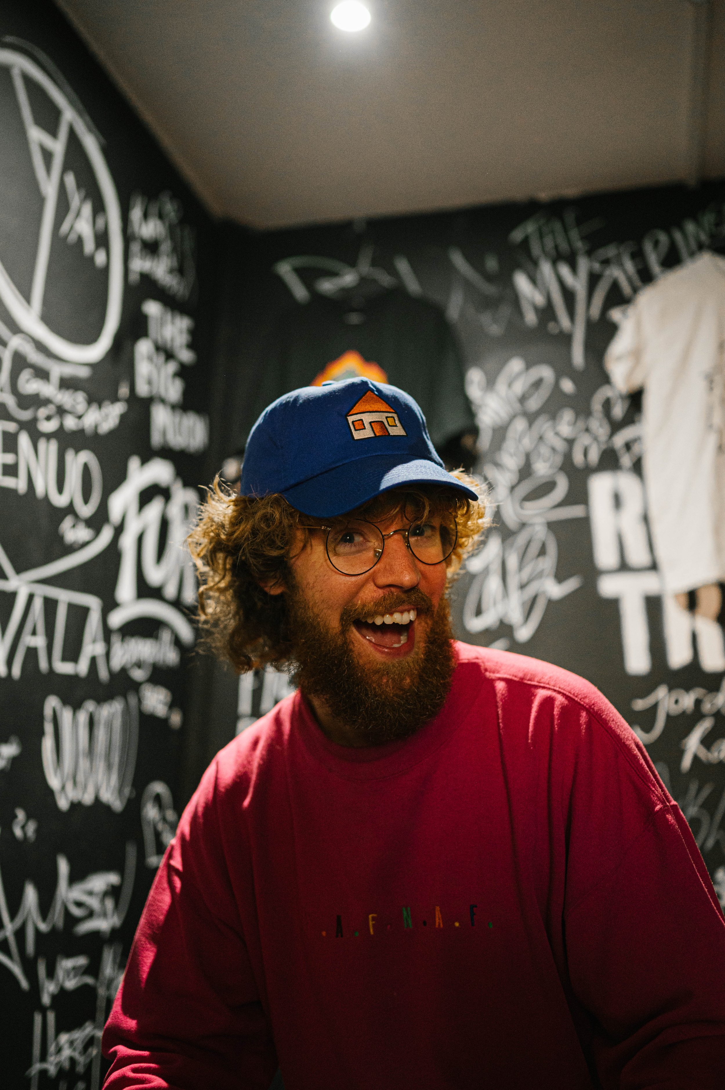 A man with curly hair and a beard, wearing glasses, a red sweatshirt, and a blue cap with a house logo, smiling in front of a black wall with white handwritten-style text and drawings.