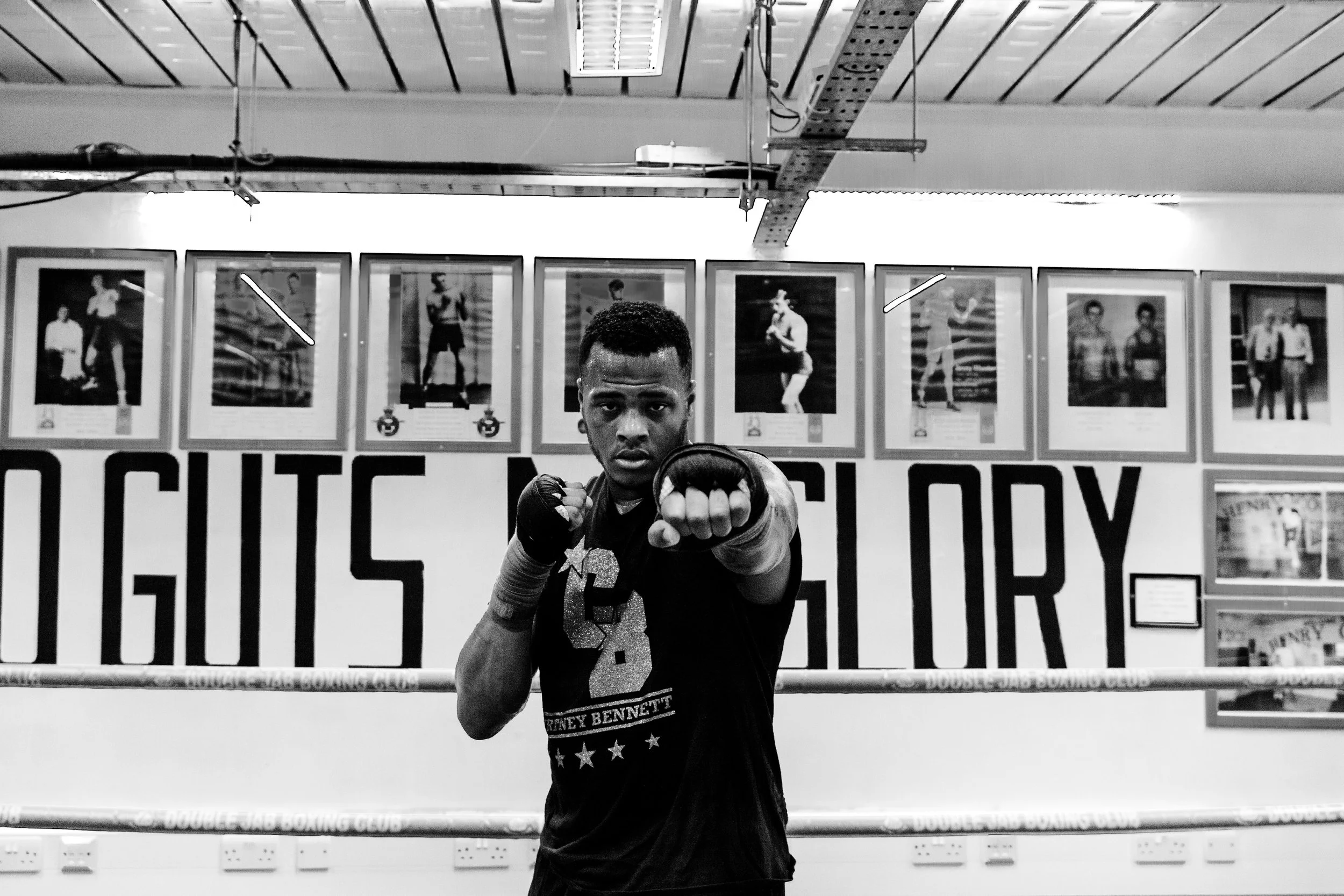 A male boxer in boxing stance inside a gym, with framed photographs of athletes on the wall behind him.