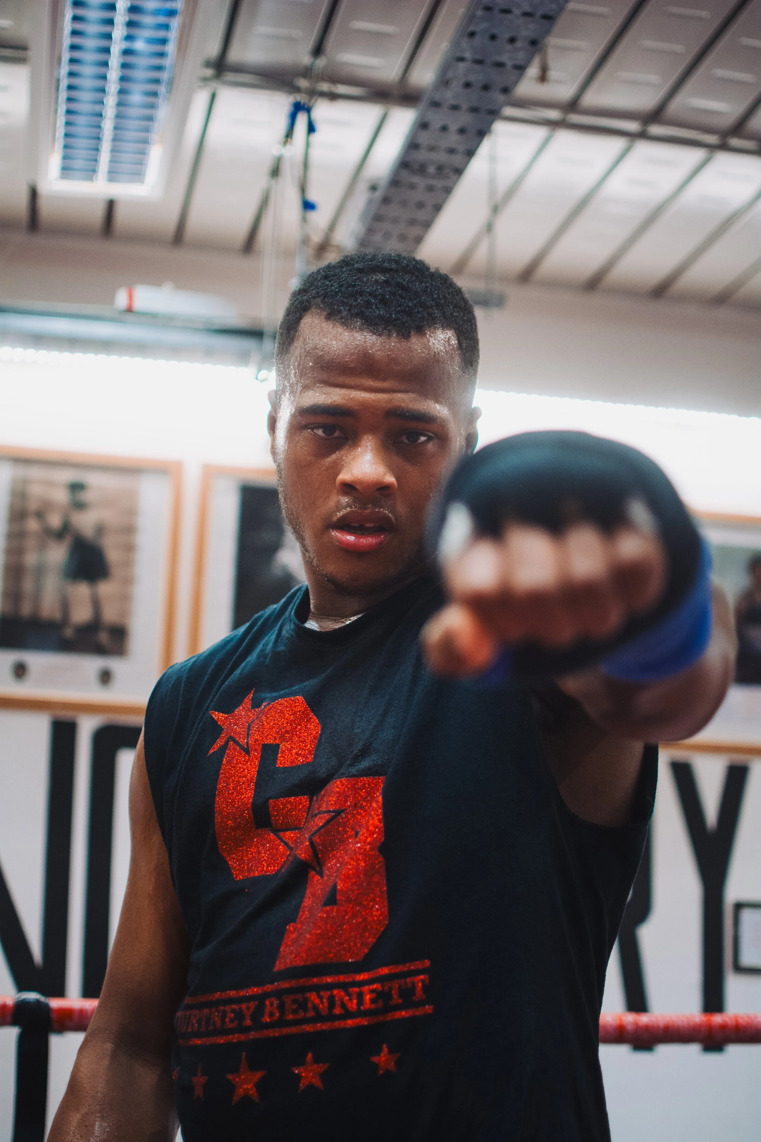 A boxer with a serious expression, wearing a black shirt with red and orange graphics, including stars and the name 'Mummey Bennett', pointing toward the camera in a gym with boxing ring ropes and photos on the wall in the background.