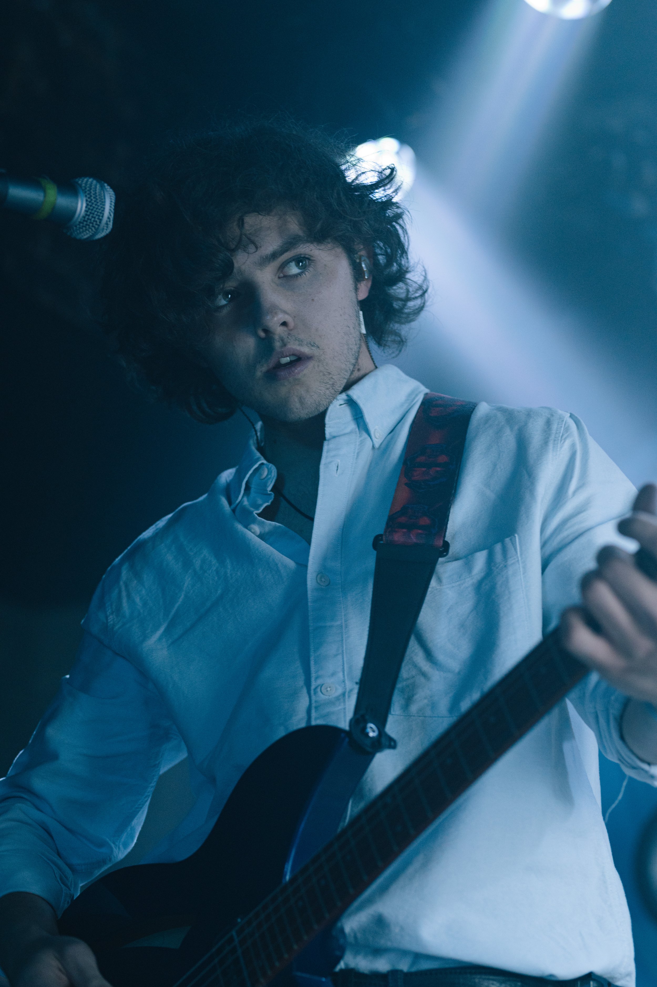 Male musician with curly hair playing guitar on stage with dramatic lighting.