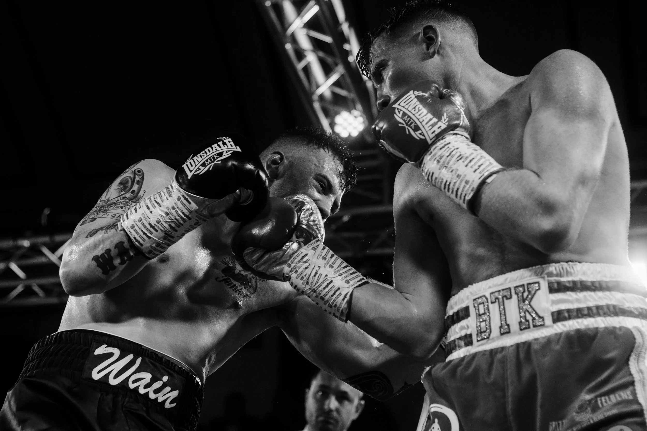 Two male boxers are engaged in a close fight inside a boxing ring. One boxer, with tattoos on his arm, is throwing a punch while the other is covering his face with gloves. The scene is intense and black-and-white.