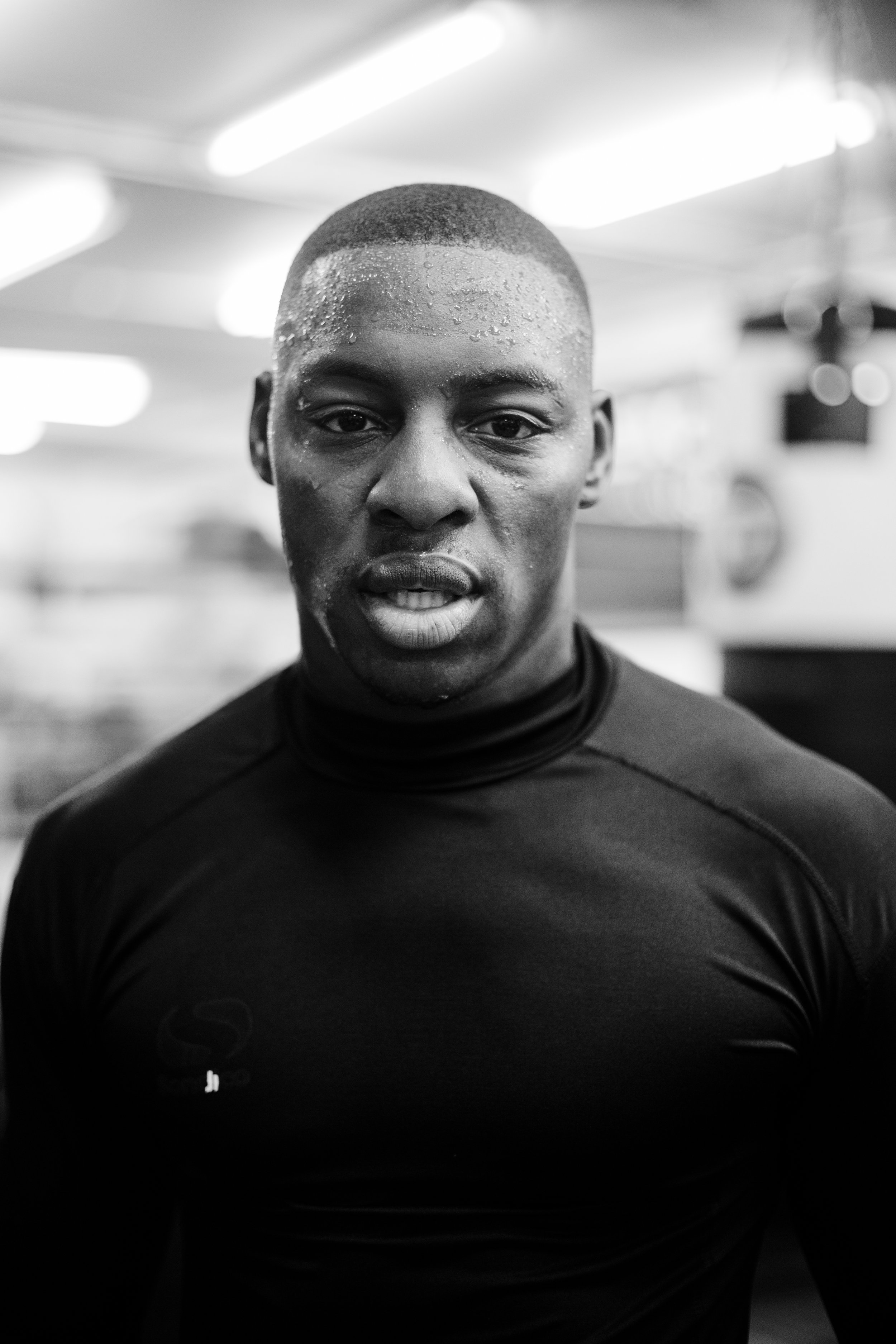 Close-up black-and-white photo of a young man with wet skin, short haircut, and athletic shirt in a gym or sports facility.