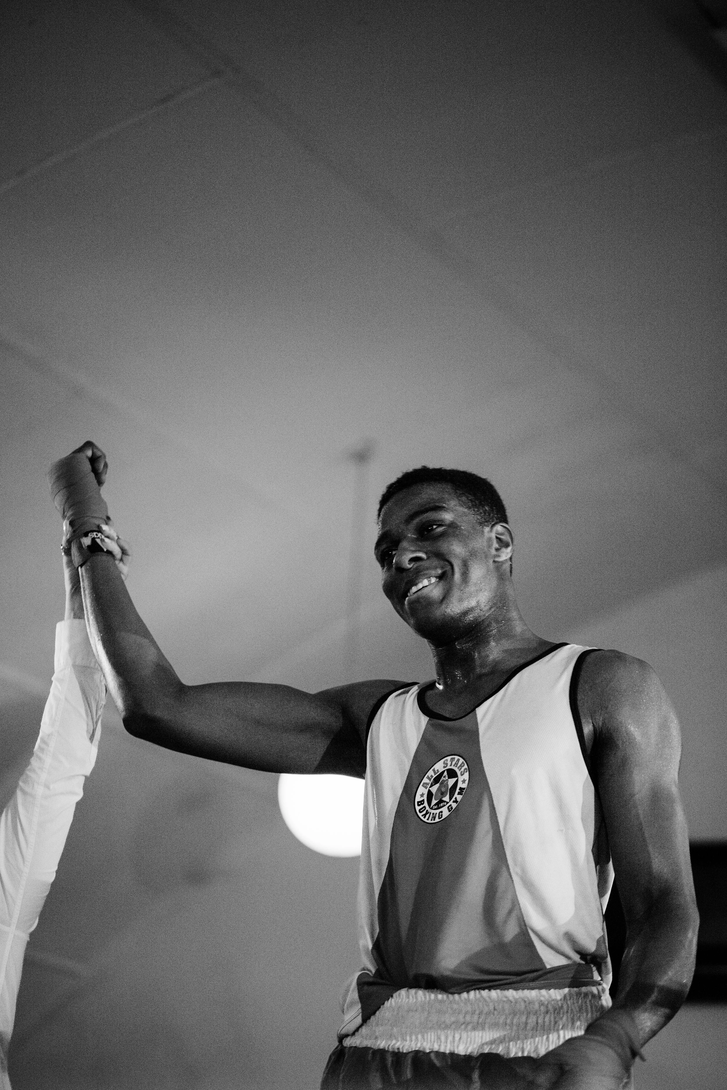 A male boxer with a smile in a boxing ring, wearing boxing attire, raising his arm in victory, with a referee extending his arm.